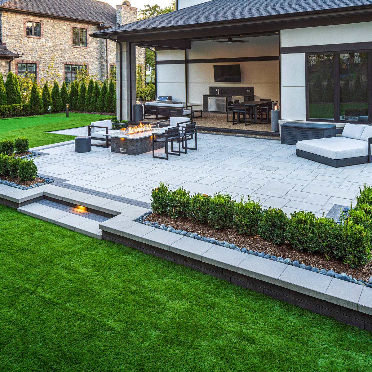 A backyard with grass and concrete pavers surrounding a fire pit.