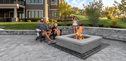 Family Sitting on Unilock Patio by Fire Pit and Person Sitting on Surrounding Retaining Wall