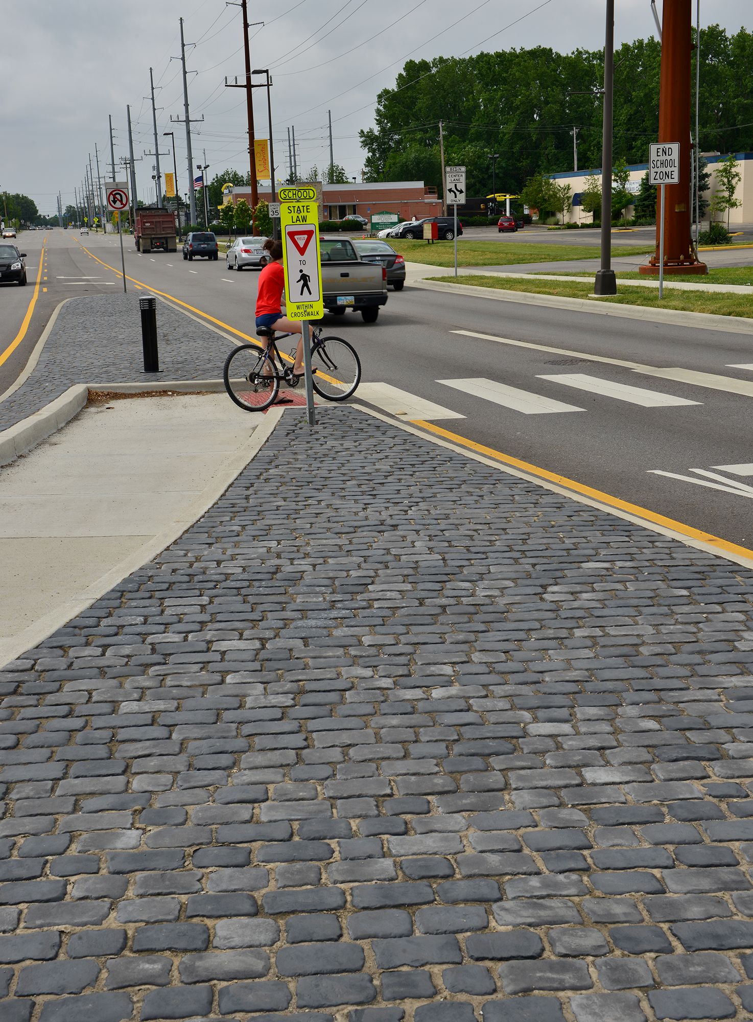 Two-toned Courtstone pavers form a triangular pattern at the edge of the sidewalk, where a cyclist prepares to cross the street.