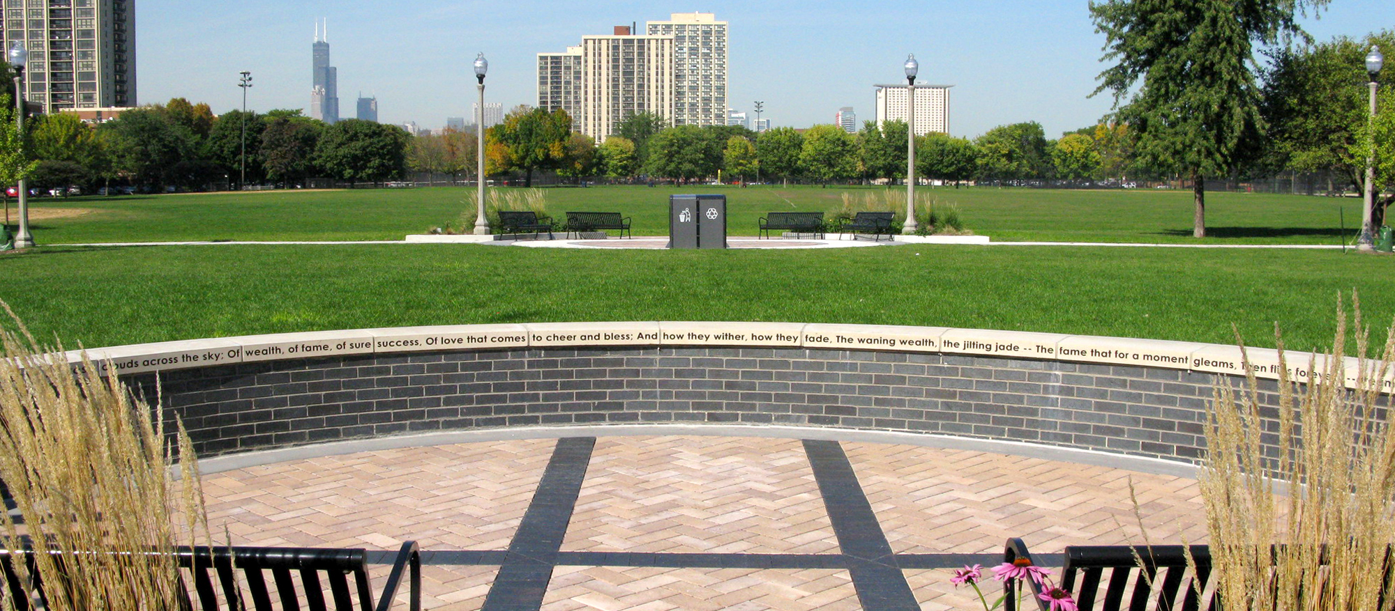 In a city park is a round plaza with contrasting lines of black Series on light Eco-Priora™ pavers ringed by a wall with a poem on its ledge.