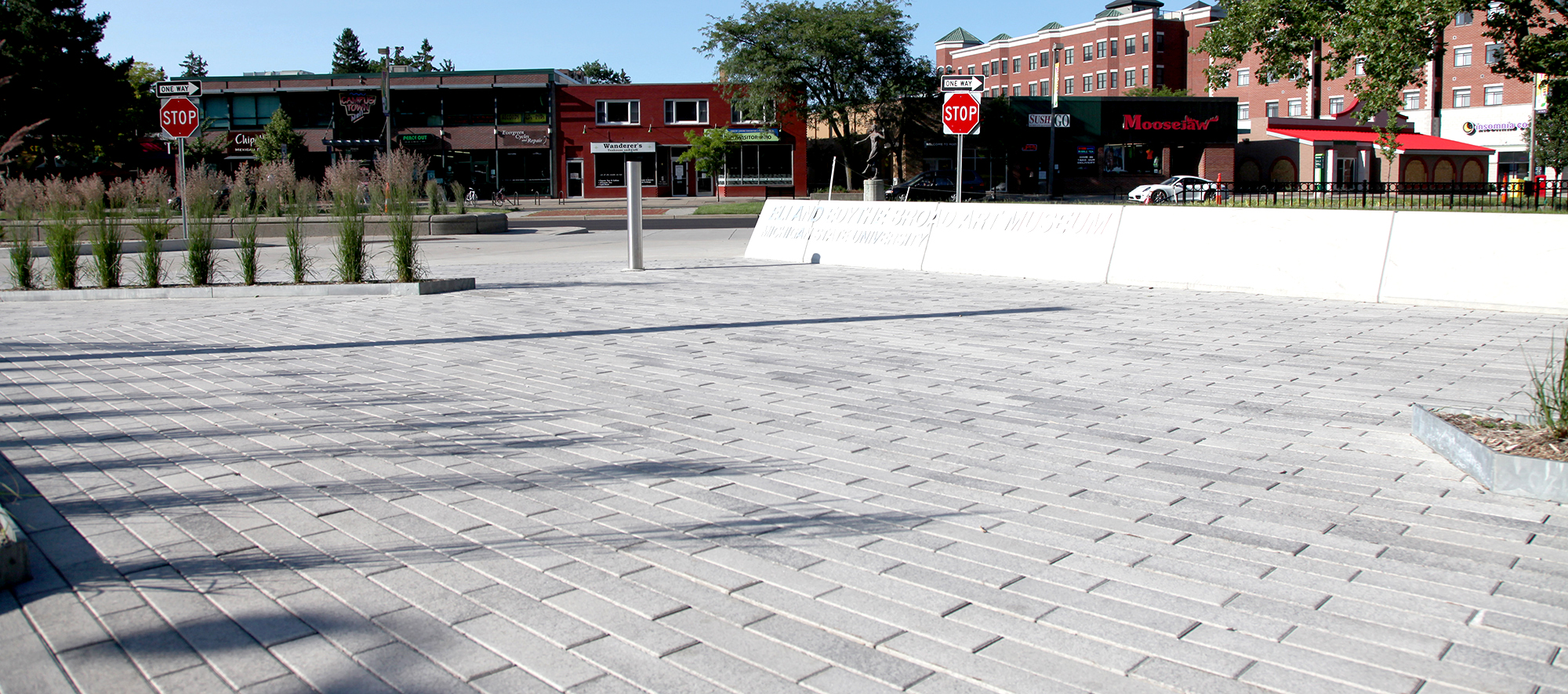 A plaza of Promenade Plank Pavers with Umbriano finish is accented with landscaping and defined by a low concrete wall.