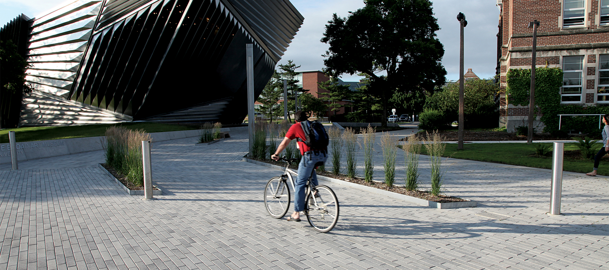 A cyclist rides on a bike path in a large area made with Promenade Plank Pavers, leading to a hexagon-shaped, striped building.