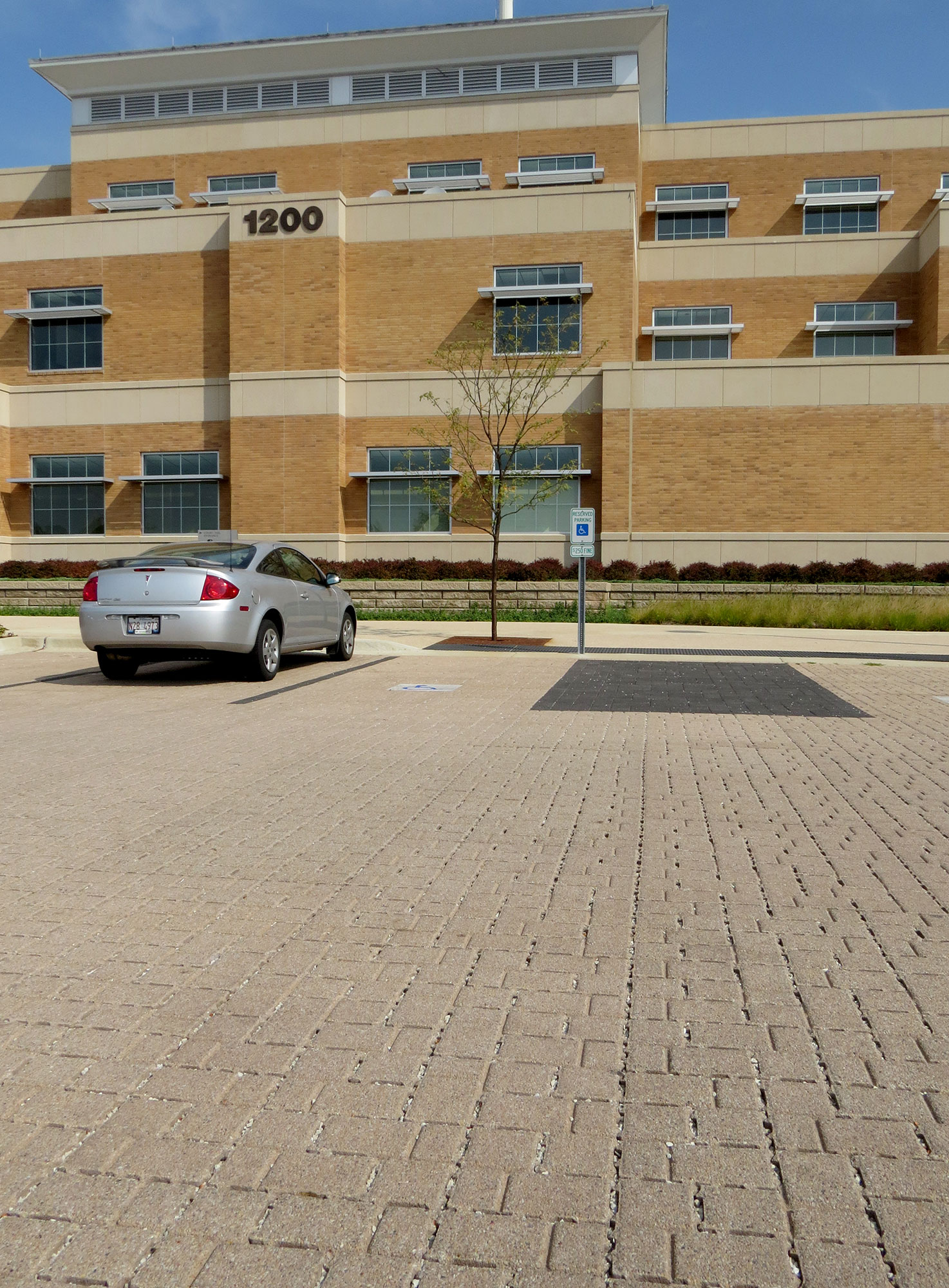 Eco-Optiloc permeable pavers in contrasting colors mark parking spots in front of a brick building with a Siena Stone walled garden.