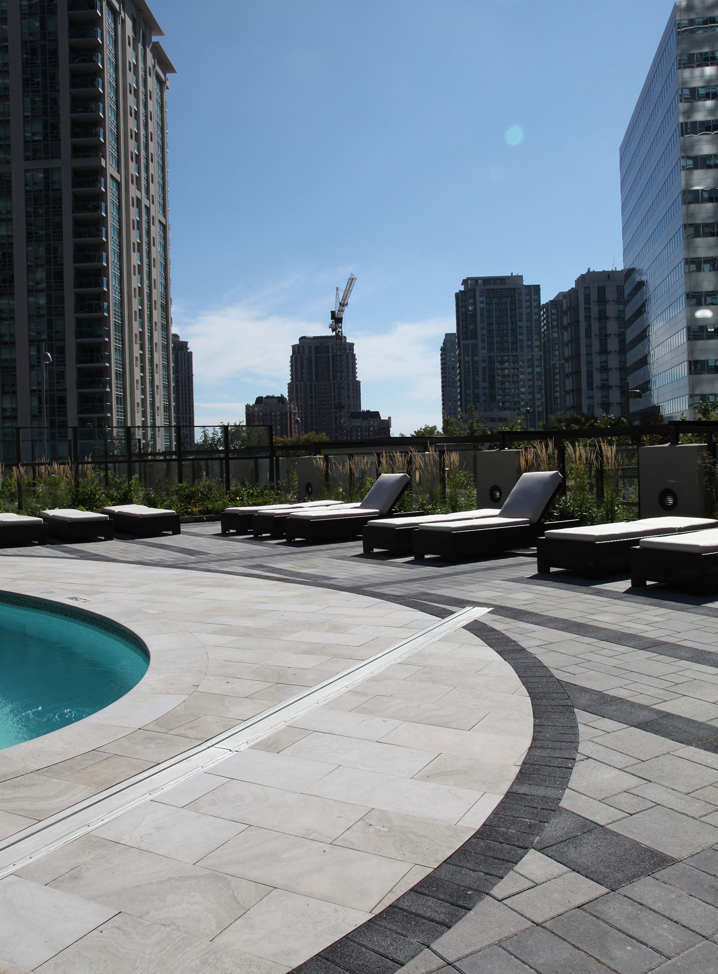 Lounge chairs wrap around the roof deck pool overlooking the cityscape, next to patterned paver rug of grey Series and black Europaver.