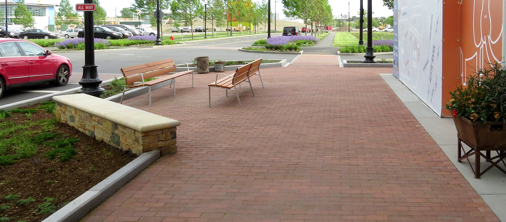 A car drives over a crosswalk beside a pedestrian area with flowers, lights, and benches; featuring Il Campo pavers in Heritage Brown.