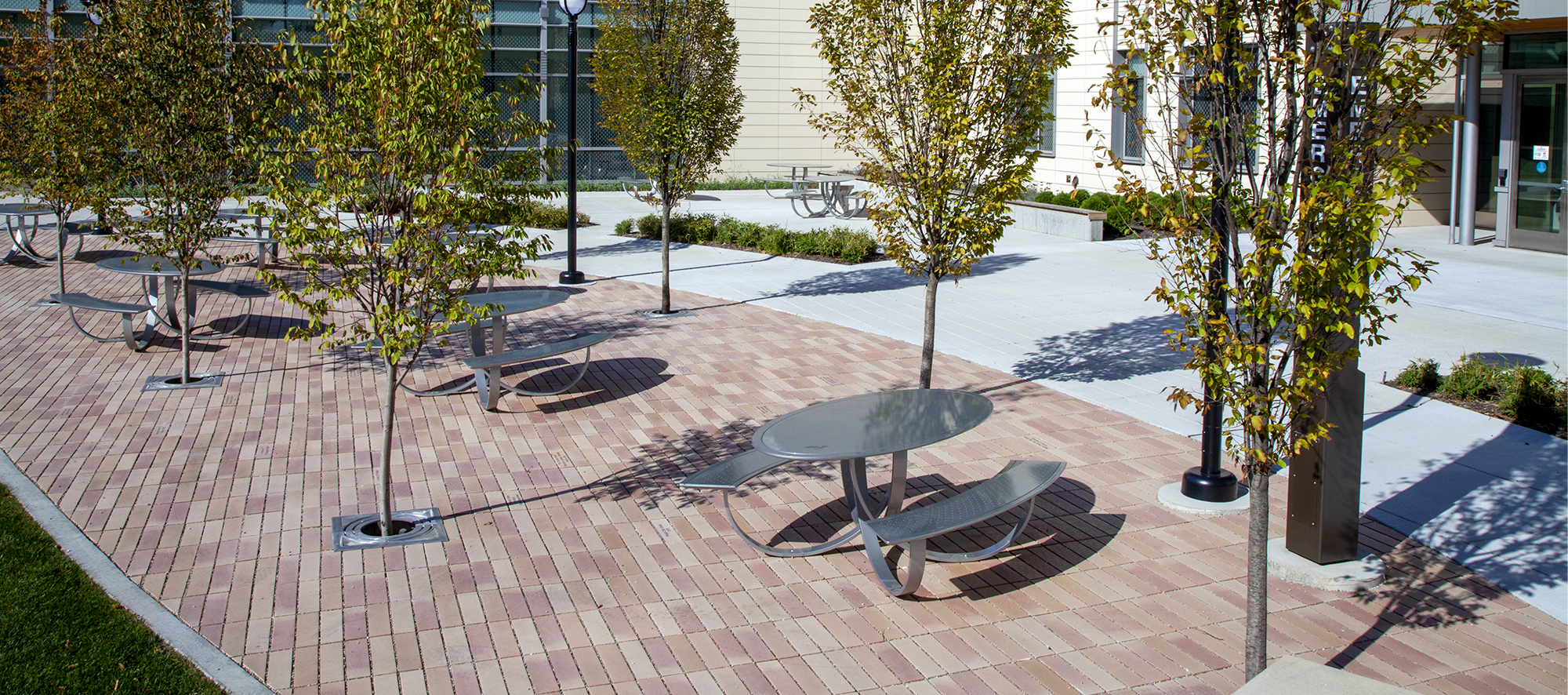 Promenade Plank pavers in a reddish-brown hue delineate a relaxing outdoor eating space, shaded by surrounding trees.