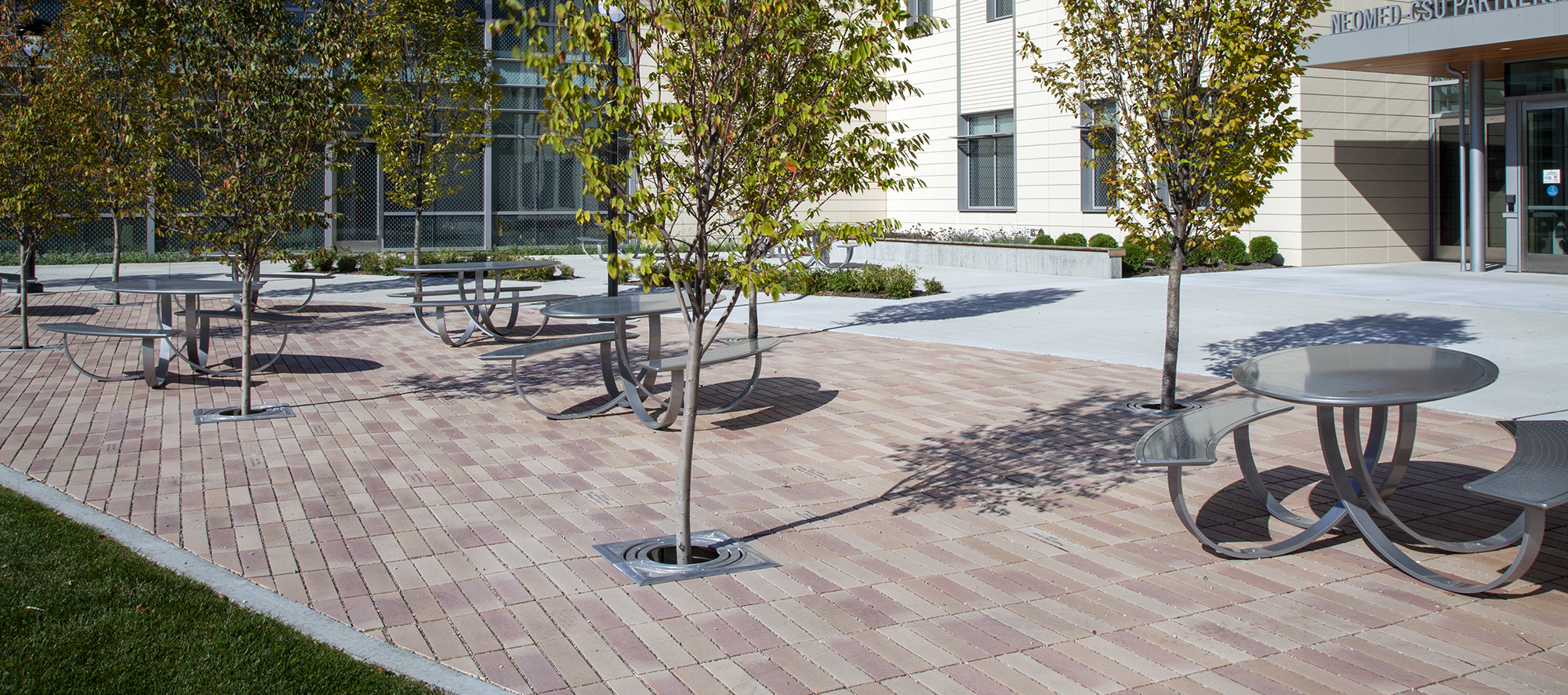 Promenade Plank pavers in a reddish-brown hue delineate an outdoor eating space, shaded by surrounding trees.