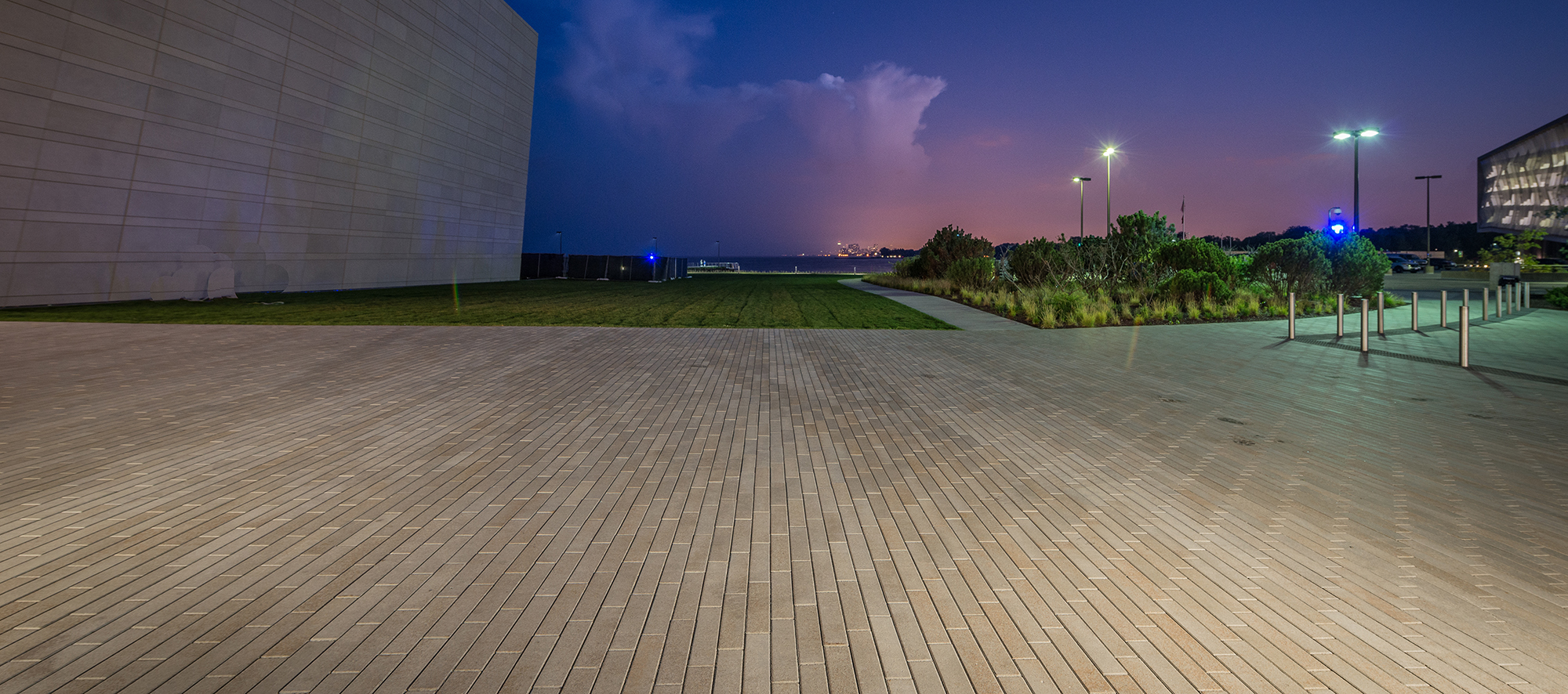 A large pedestrian area of Promenade plank pavers bordered by landscaping and a parking lot is lit by streetlights and the purple evening sky.