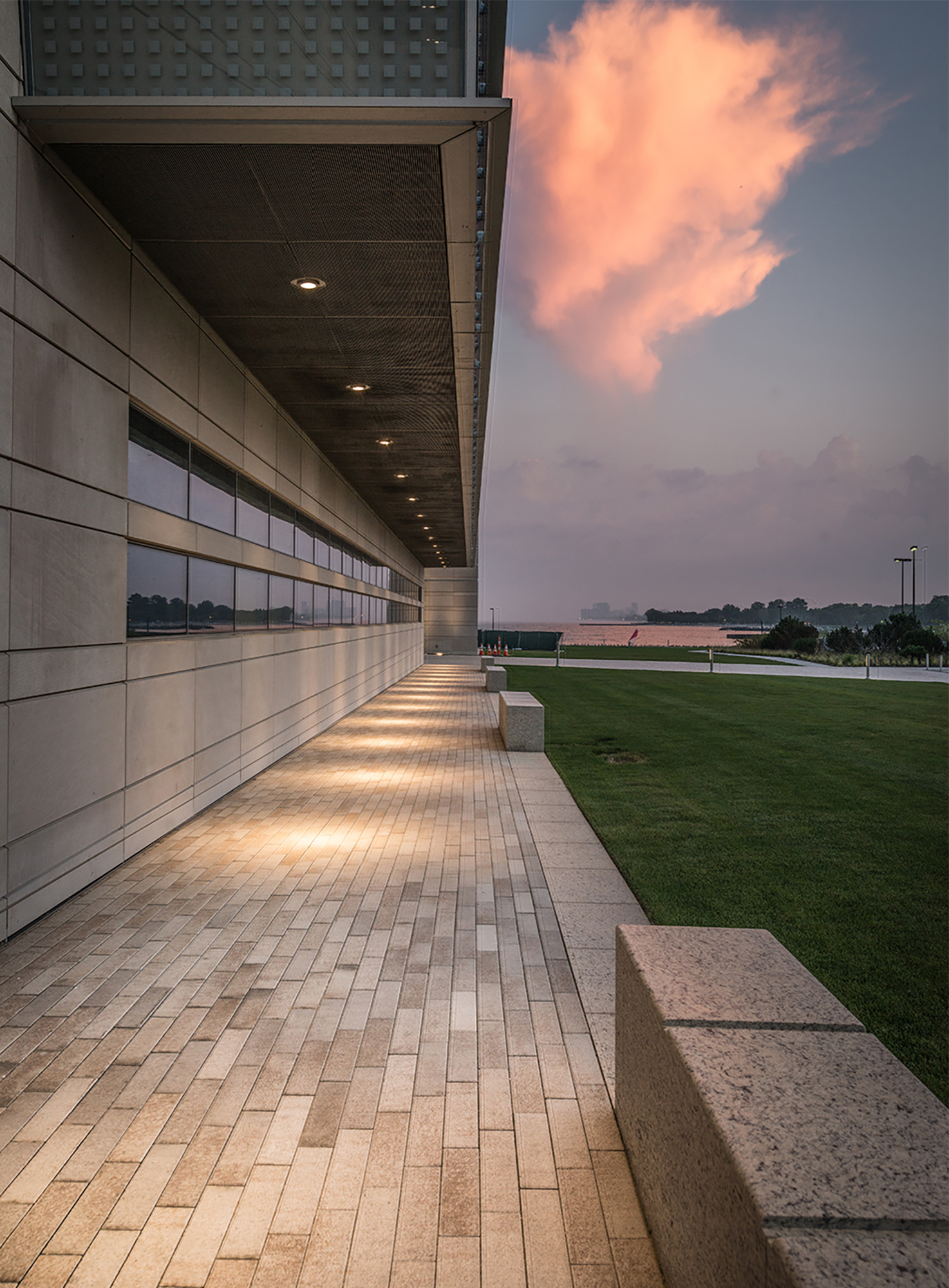 The side of a building with a path of Promenade Plank pavers in the color summer wheat, benches and grass set against the early morning sky.