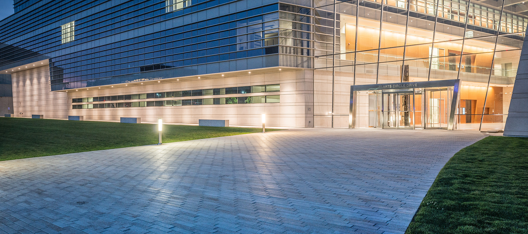 An entryway of a glass and steel building with outdoor lighting has grassy areas and a large path paved with Promenade plank.