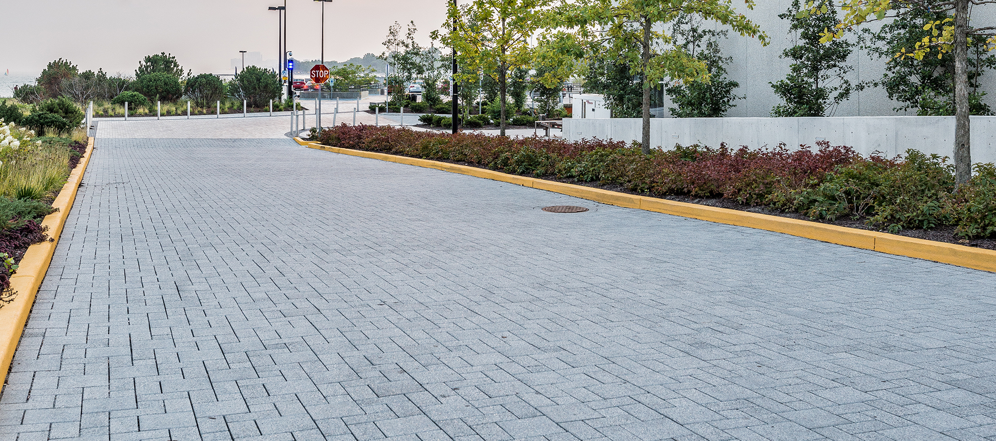 A road leading to a parking lot is paved with varying shapes of Eco-Priora permeable paver in mineral ice grey and bordered by trees and landscaping.
