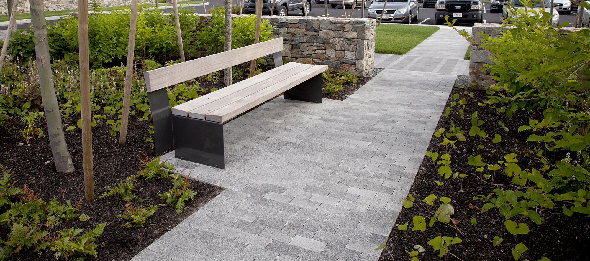 A park bench is surrounded by simple shrubs, trees, a retaining wall, and a white-grey Umbriano paver walkway toward the parking lot.