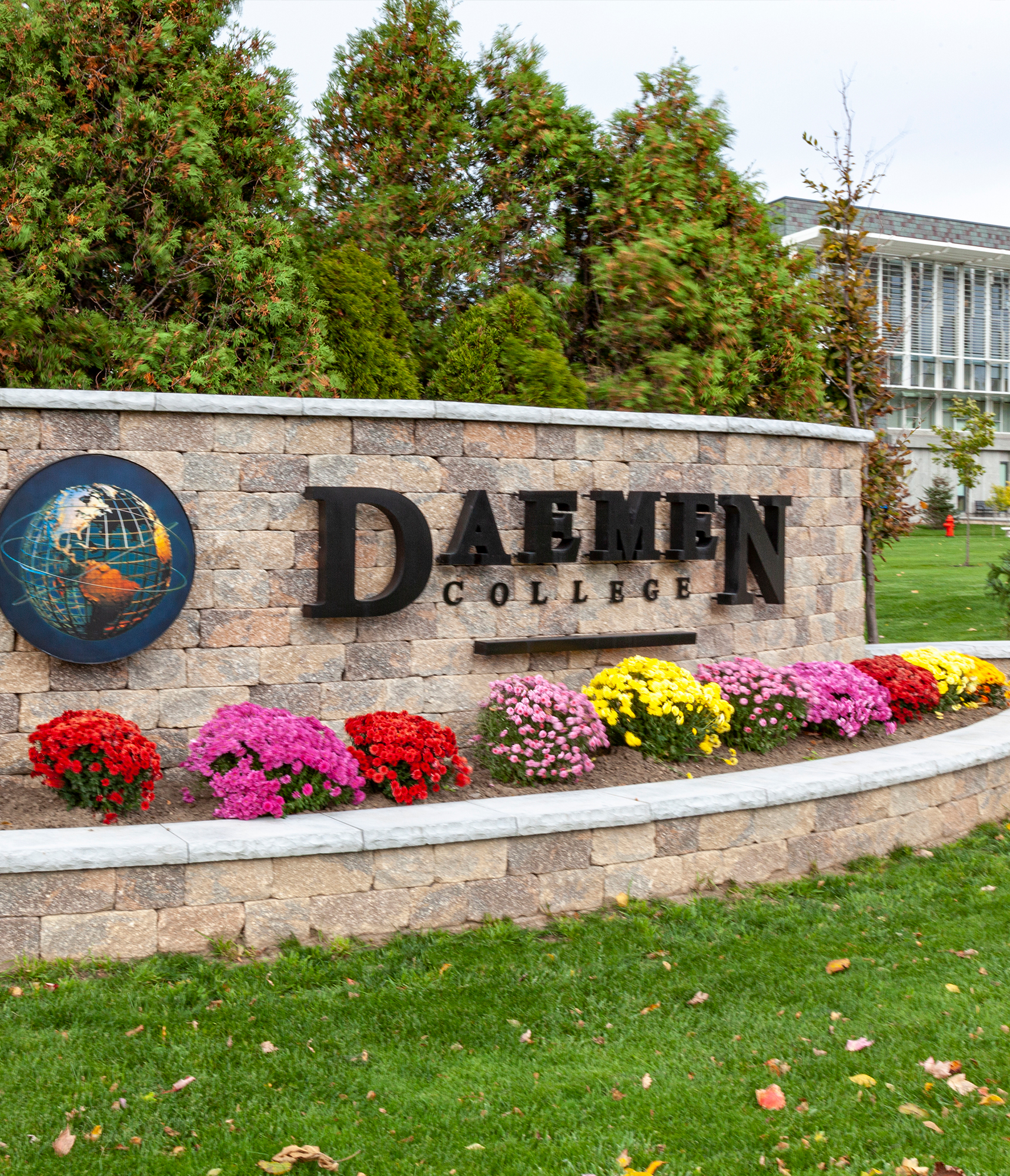 With a parking lot and building in the background, a 2 Level Estate Wall on the lawn holds the logo for Daemen College, flowers, and trees.