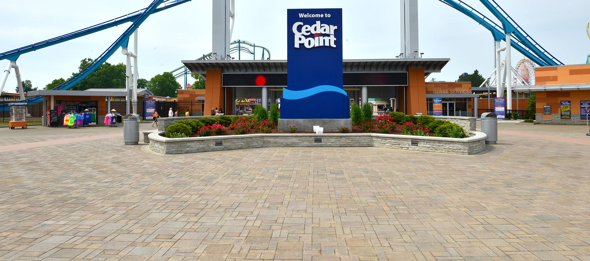 A large rollercoaster sits behind a retaining wall with lush softscaping featuring a large Cedar Point sign, over beige Il Campo pavers.
