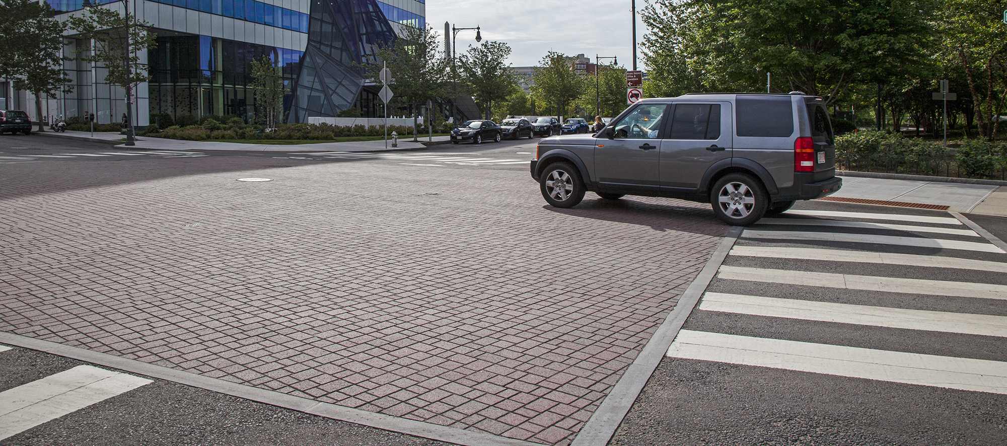 A vehicle, cyclist and pedestrian cross through the intersection, passing over a square paver bed of red Optiloc pavers.
