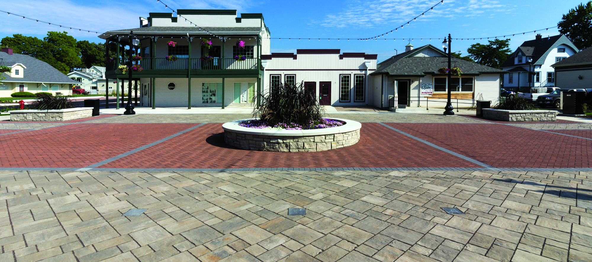 A pedestrian area in front of vintage buildings features contrasting colors and sizes of Unilock pavers, a raised garden, and string lights.