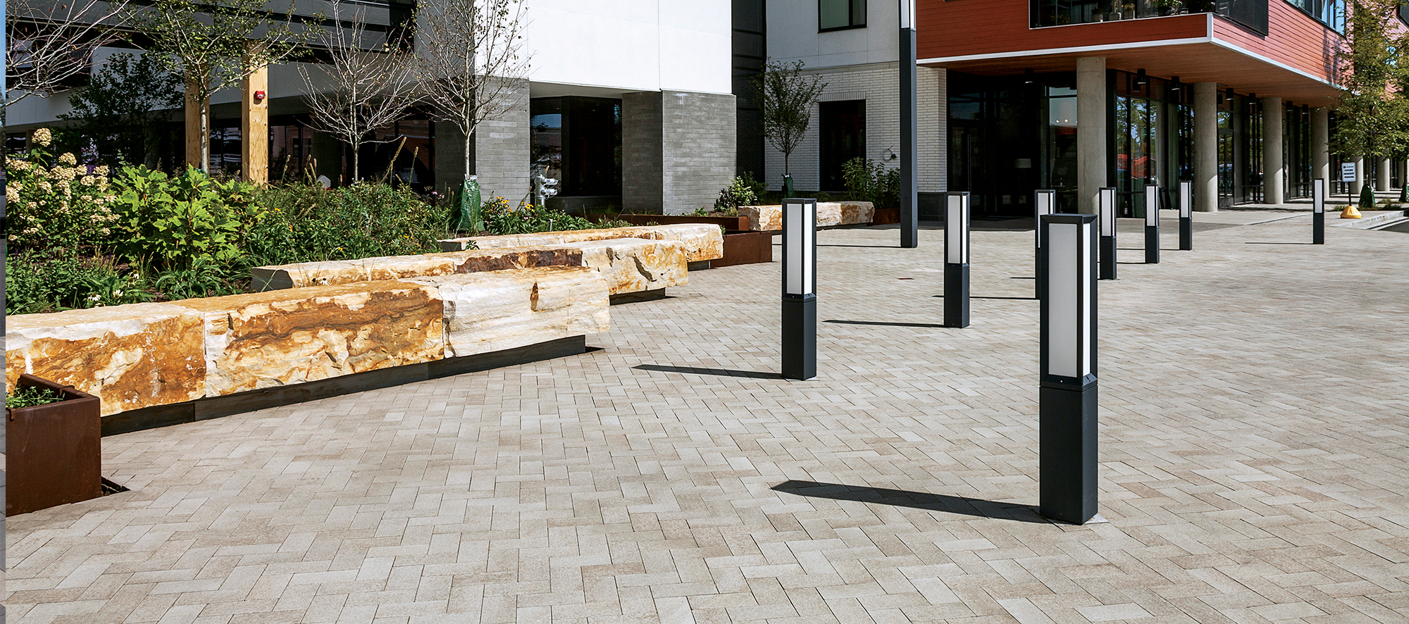 Decorative light posts cast shadows over beige Umbriano pavers, with a retaining wall and garden bed bordering the outside of the complex.