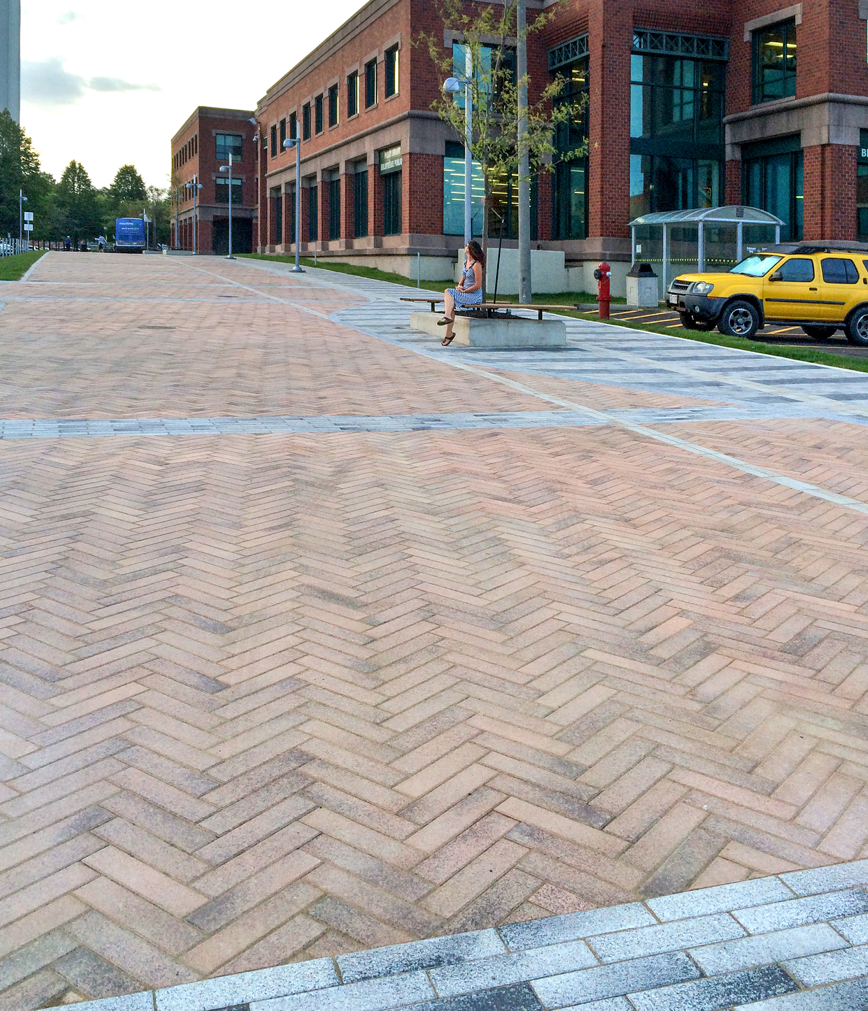  A woman sits on a bench in  pedestrian area paved with warm tones of Umbriano pavers in a zig-zag pattern contrasted with lines of grey.