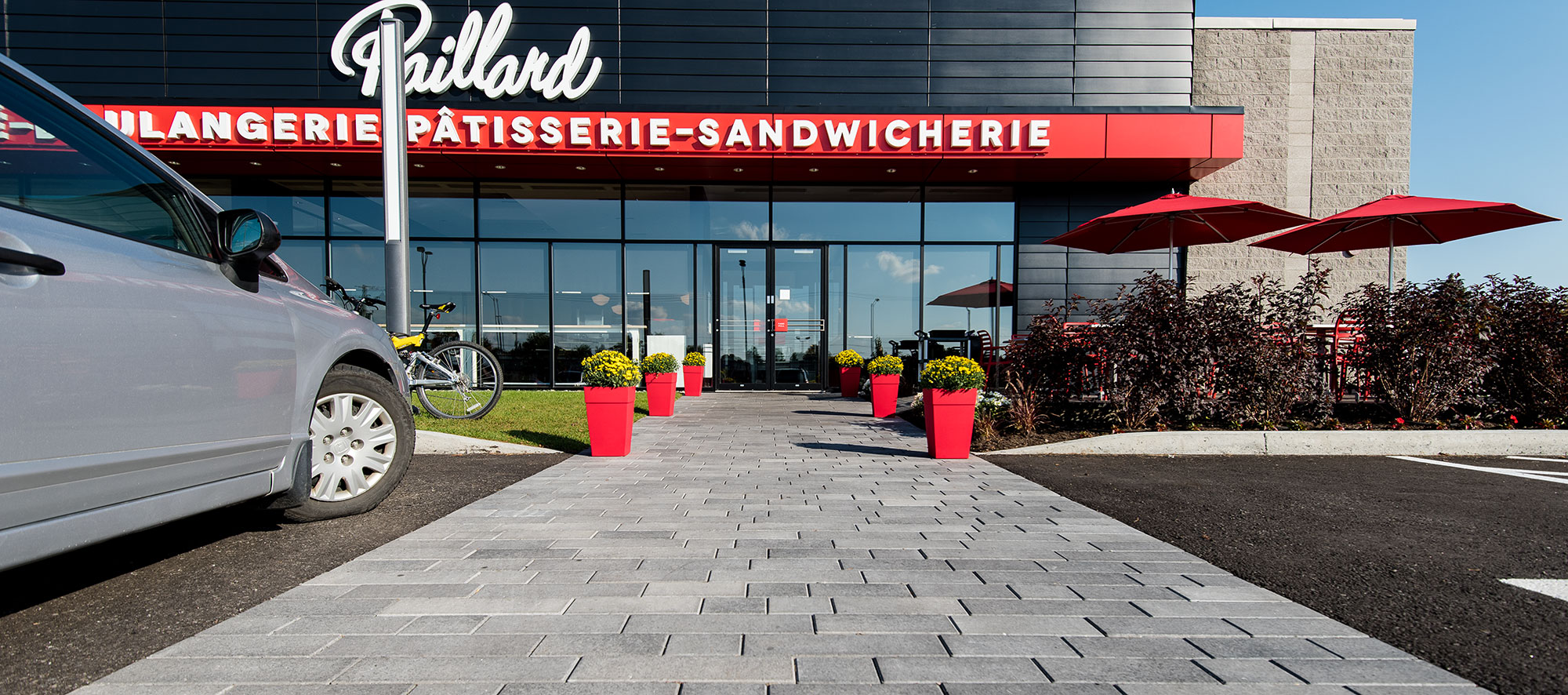 A café entrance includes gardens, red umbrellas, flowers in red planters, and a walkway of Artline pavers with a mottled Umbriano finish.