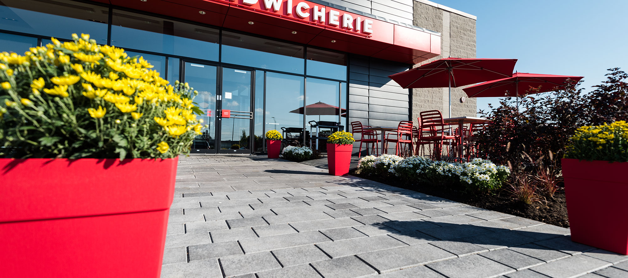 Artline pavers, landscaping and an outdoor dining area with umbrellas and planters matching the red theme, lead to Boulangerie Paillard.