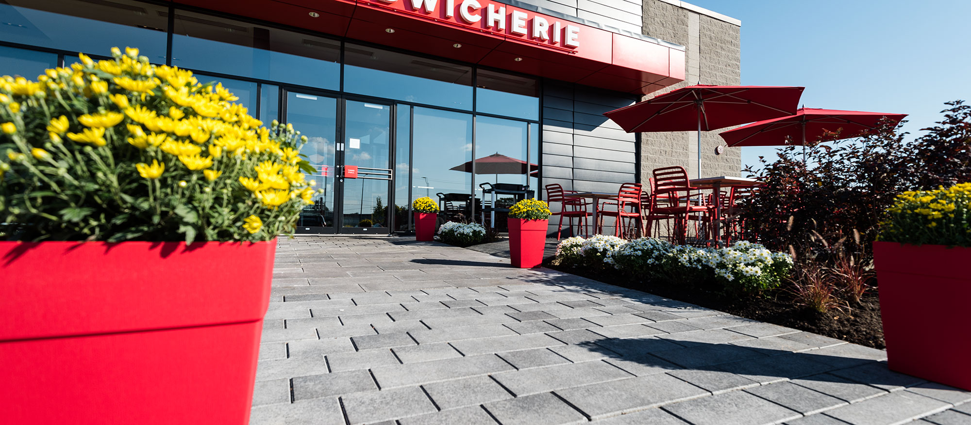 Artline pavers, landscaping and  an outdoor dining area with umbrellas and planters matching the red theme, lead to  Boulangerie Paillard.