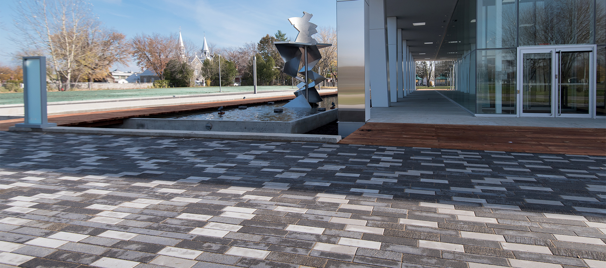 Tri-colored Il Campo pavers make up a grand walkway to the entrance of the building, where a metal art piece sits within a water fountain.