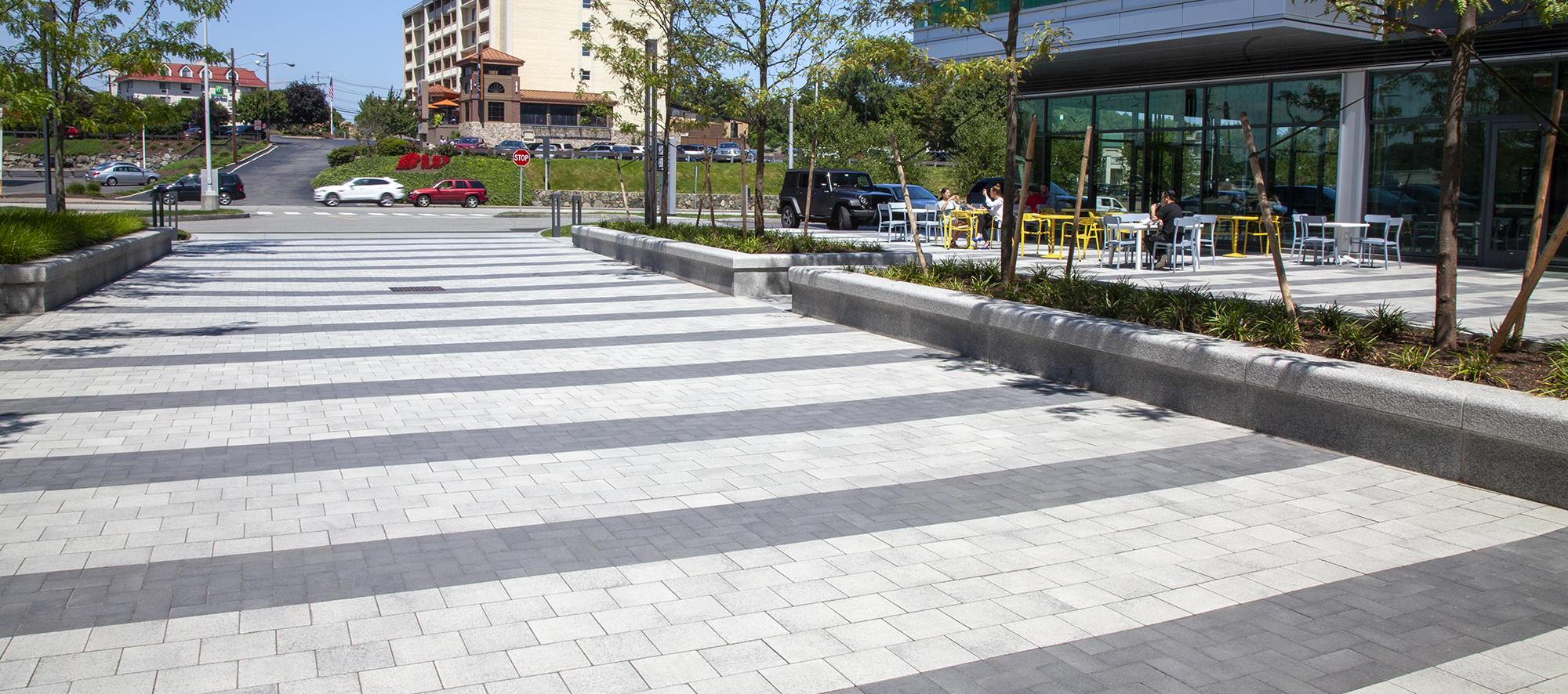 A striped pedestrian path made with contrasting shades of Umbriano pavers is bordered by a roadway and businesses with outdoor seating.