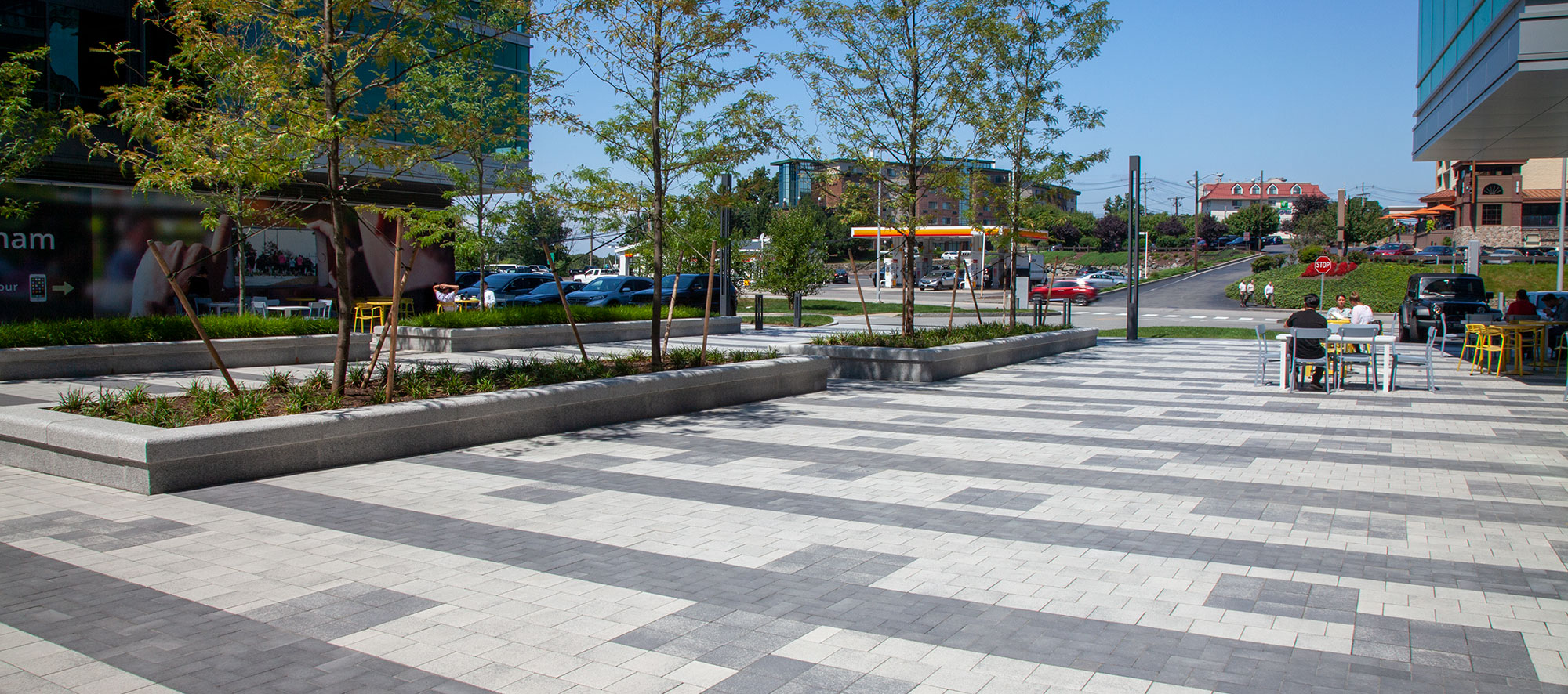 A plaza made from Unilock Umbriano pavers in a geometric pattern is lined by trees, gardens and seating areas.