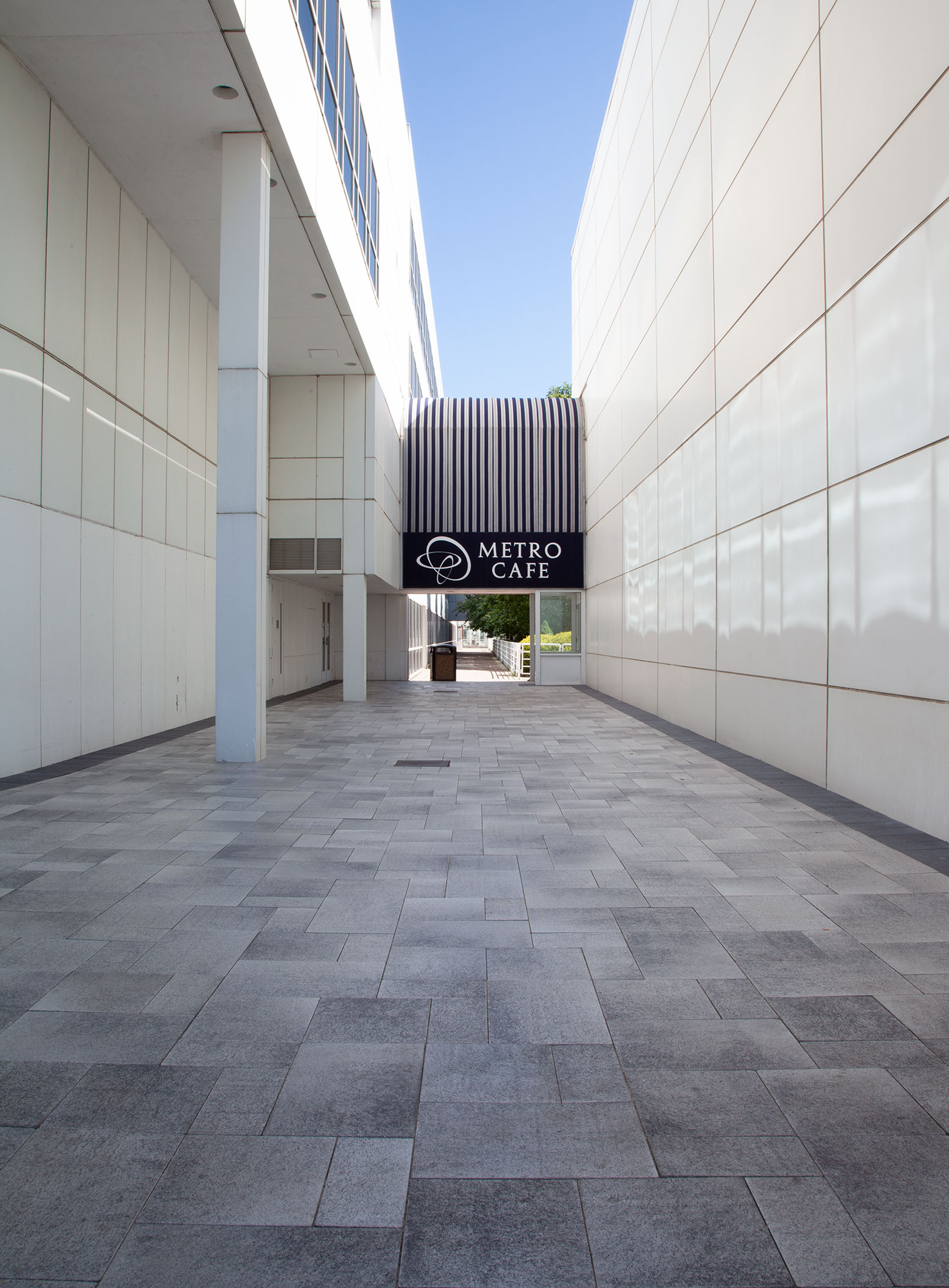 Between two white buildings, a path of grey Umbriano pavers in varying sizes and shades leads to the patio entrance of the Metro Café.