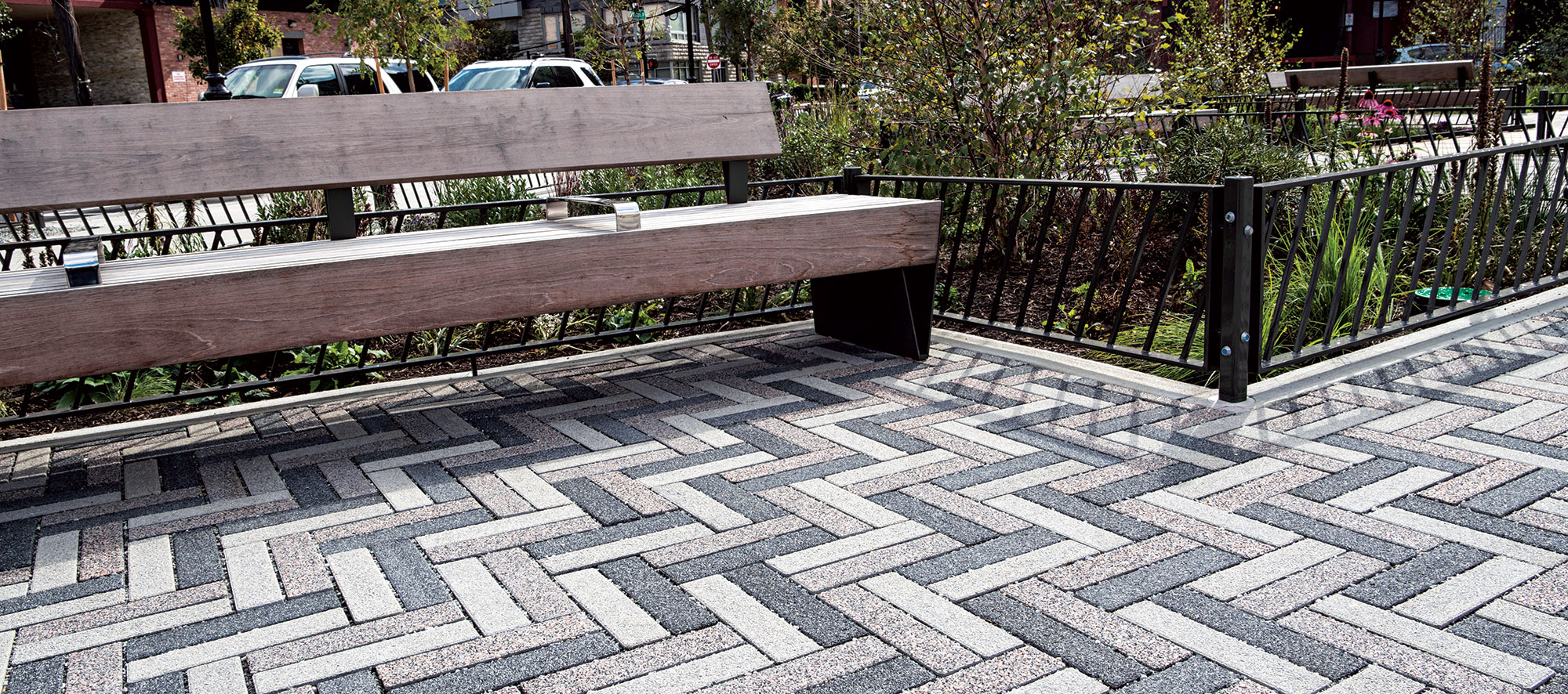 Tri-colored Eco-Promenade pavers glimmer next to a distinct wooden park bench, with an adjacent green island outlined by metal fencing.