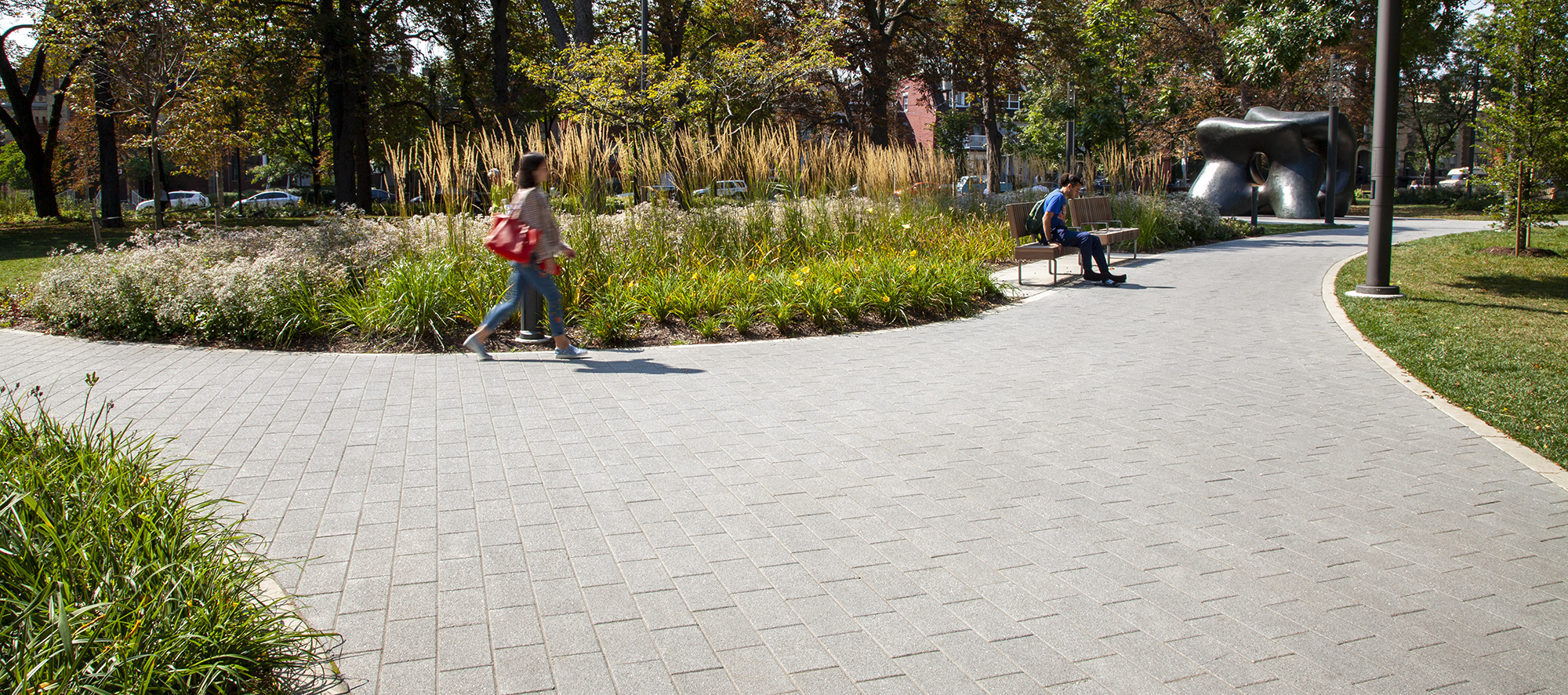A visitor strolls past a garden bed while a pedestrian sits on a bench, with the backdrop of a bronze sculpture atop grey Series pavers.