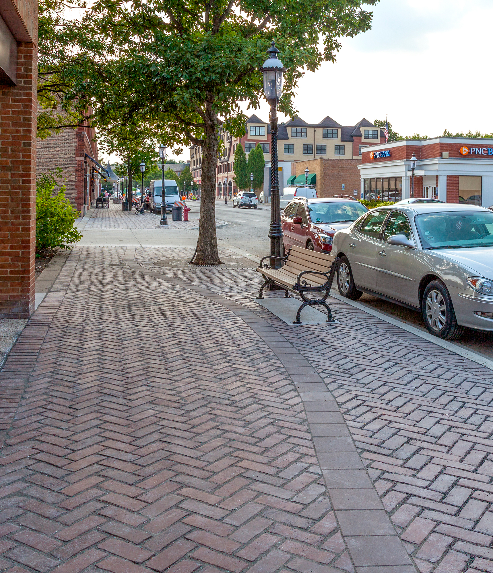 A gently curving line of pavers disrupts the zigzagging Town Hall pavers on the sidewalk with a bench, streetlight and trees.