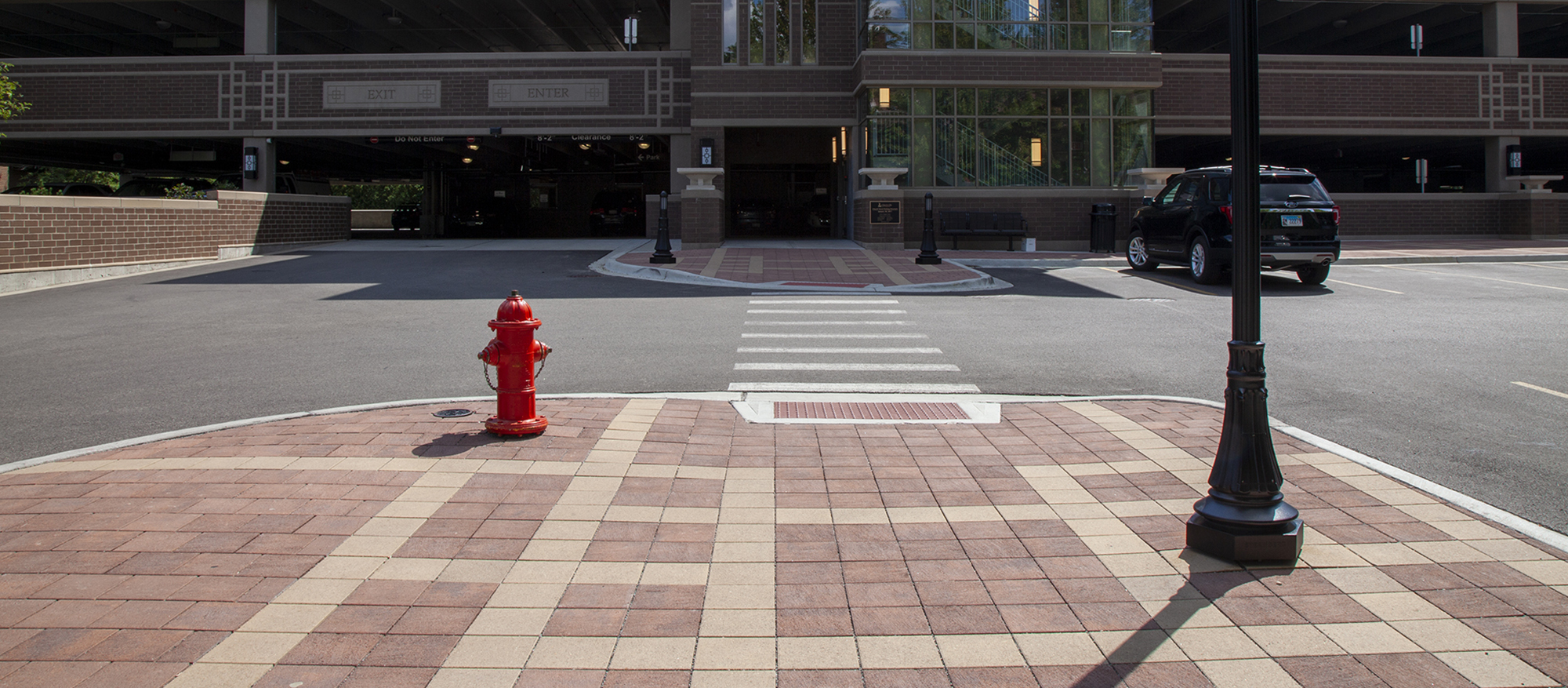 A car is  parked in front of a parking garage in an outdoor lot with raised curbs of red and beige Eco-Priora pavers, lights and a hydrant.
