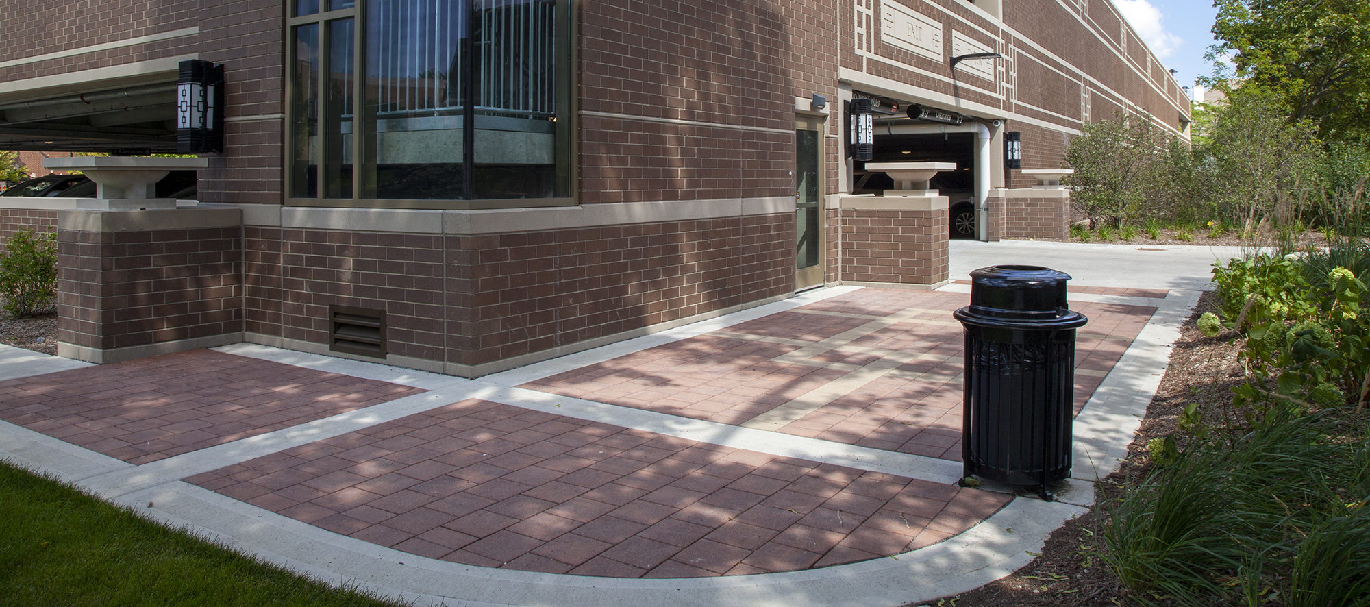 Red and beige Eco-Priora pavers continue around the corner of a red brick parking garage with a garbage can on the side.