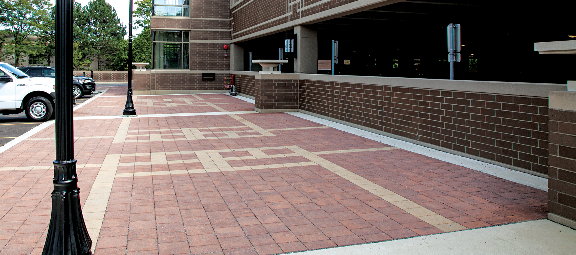 A sidewalk of red and beige patterned Eco-Priora pavers between cars parked in an outdoor lot and the wall of an enclosed parking garage.