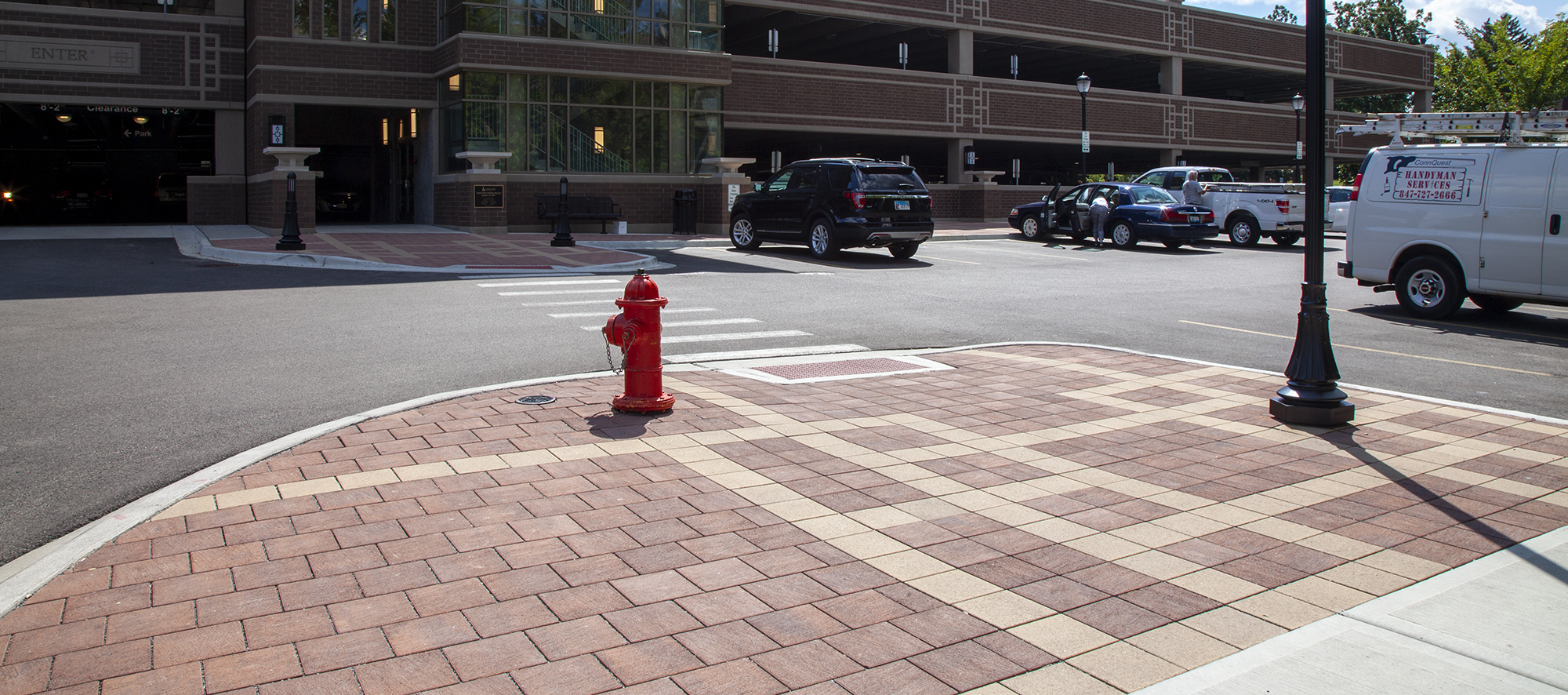 Cars are parked in front of a parking garage in an outdoor lot with raised curbs of red and beige Eco-Priora pavers, lights and a hydrant.