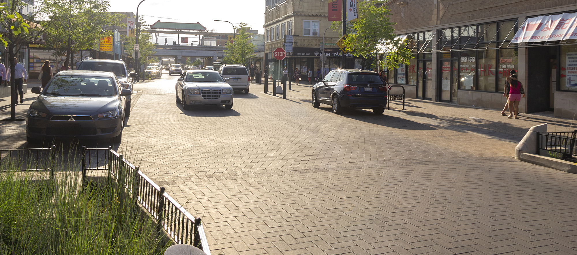 Unilock Eco-Priora pave Argyle Street roadway and sidewalk. People walk on the treelined sidewalk as cars park and drive on the road.