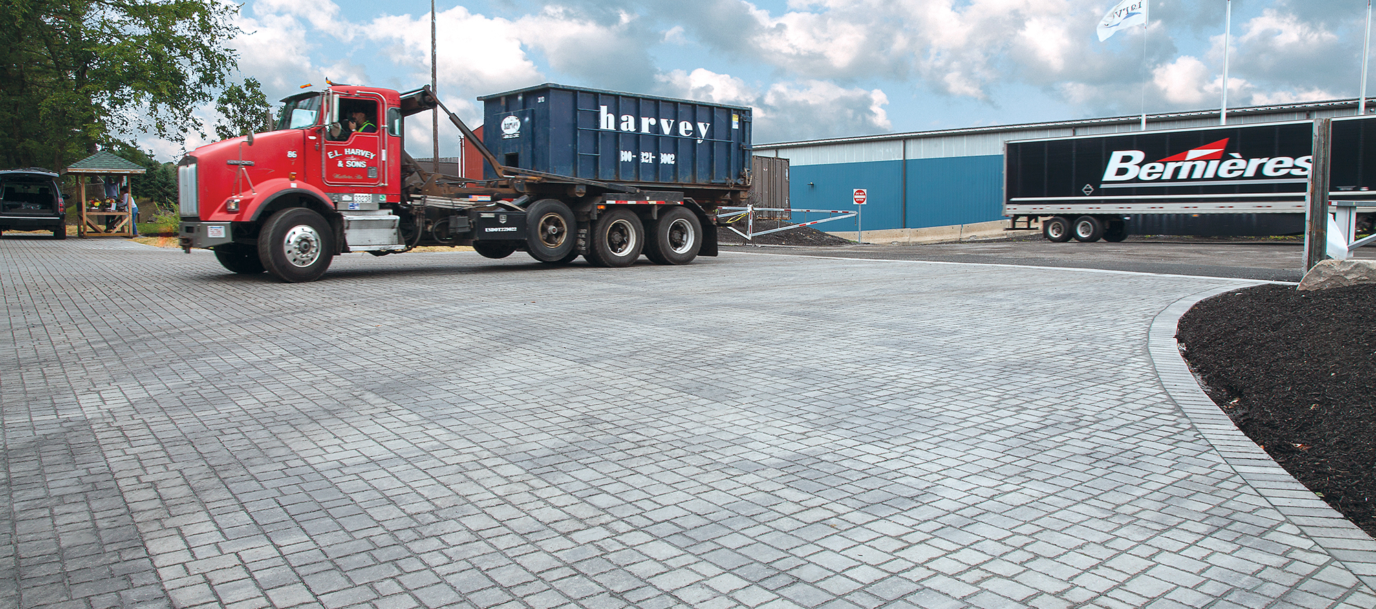 An E.L. Harvey & Sons truck exits the industrial site, driving over a bed of grey Optiloc pavers arranged in a random pattern.