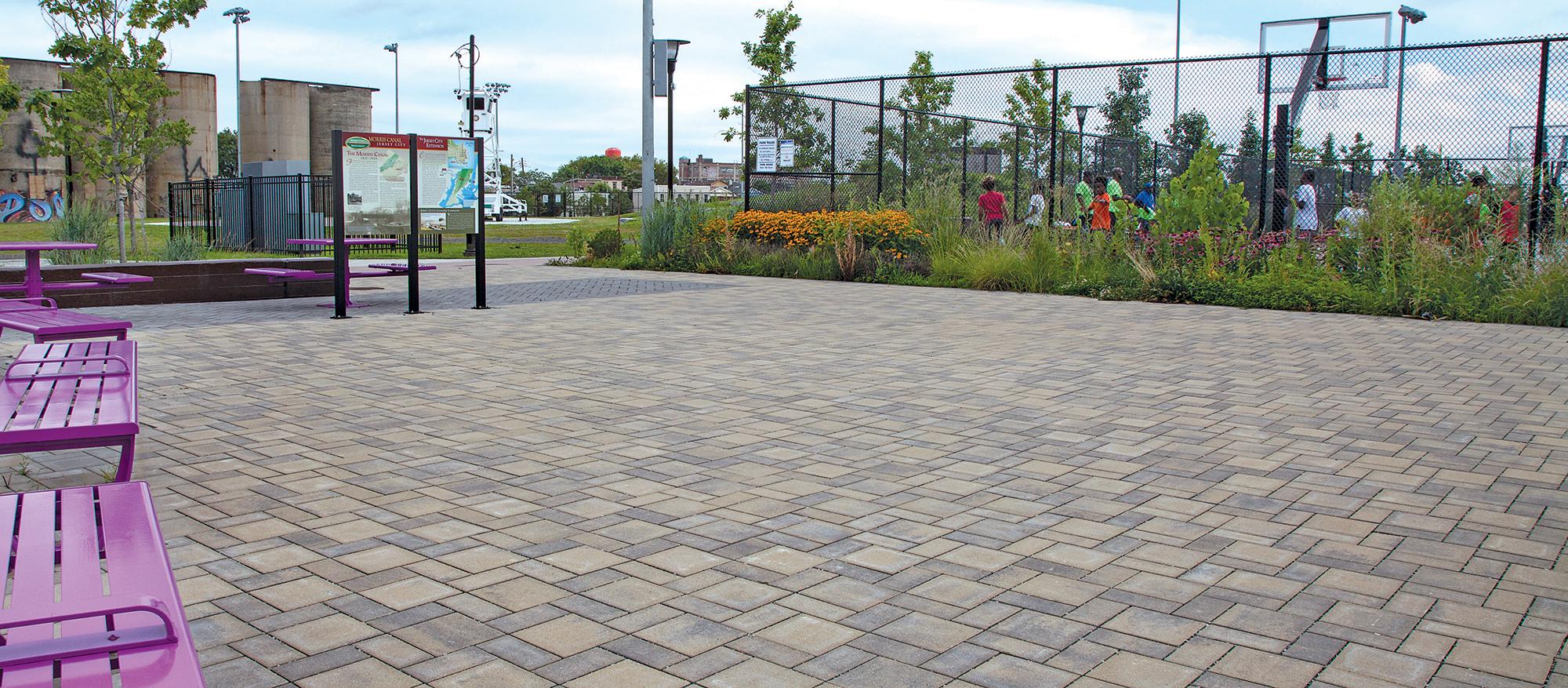 A parkette with Eco-Priora permeable pavers with bright benches and picnic tables next to a garden and basketball court.
