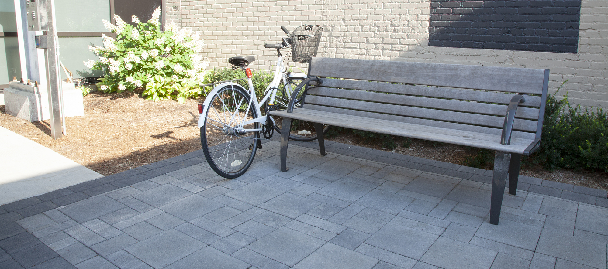 A bicycle and a bench sit on Unilock Il Campo pavers in different shapes and shades of grey, Surrounded by paved gardens and a wall.
