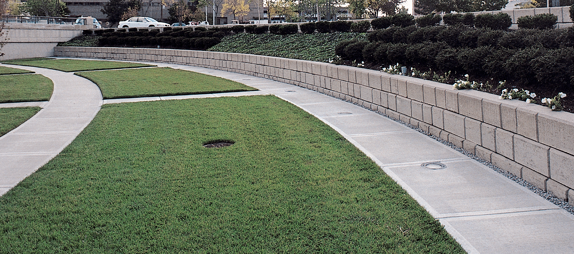 A DuraHold2 retaining wall gently curves, running parallel with the concrete pathway and green patches while containing its own softscaping.