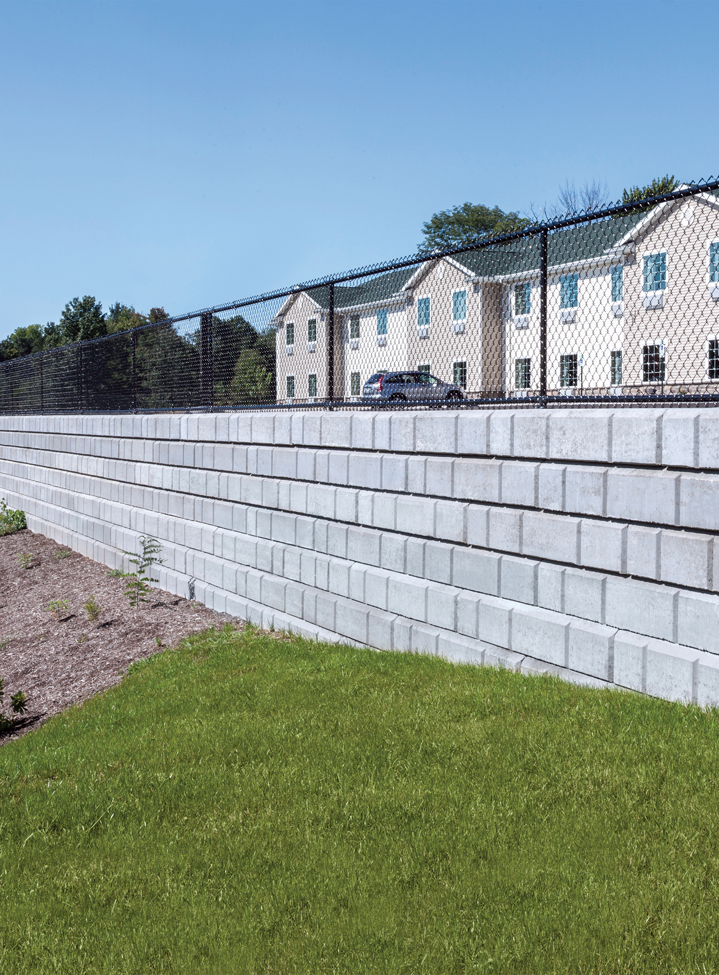A chain-link fence lines the top of the Dura-Hold retaining wall, defining the upper parking area from the lower garden island.