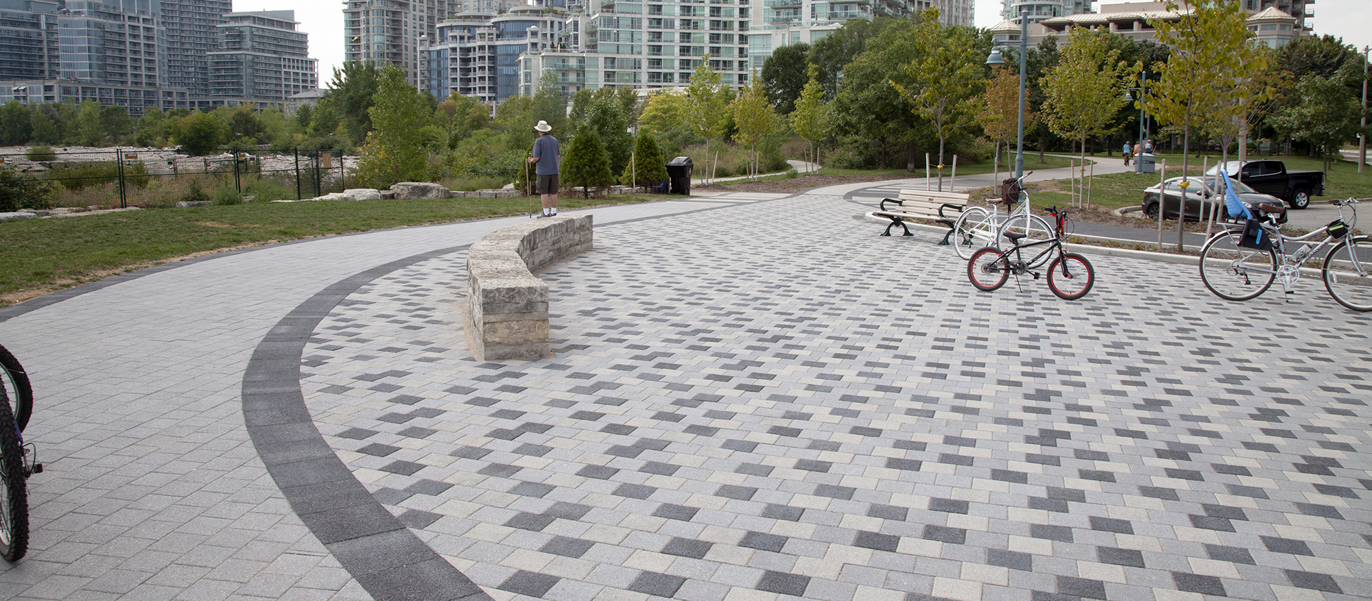 Bikes sit parked on a checkerboard paver rug of tri-colored Series pavers, as a visitor stands atop a low seat wall nearby.