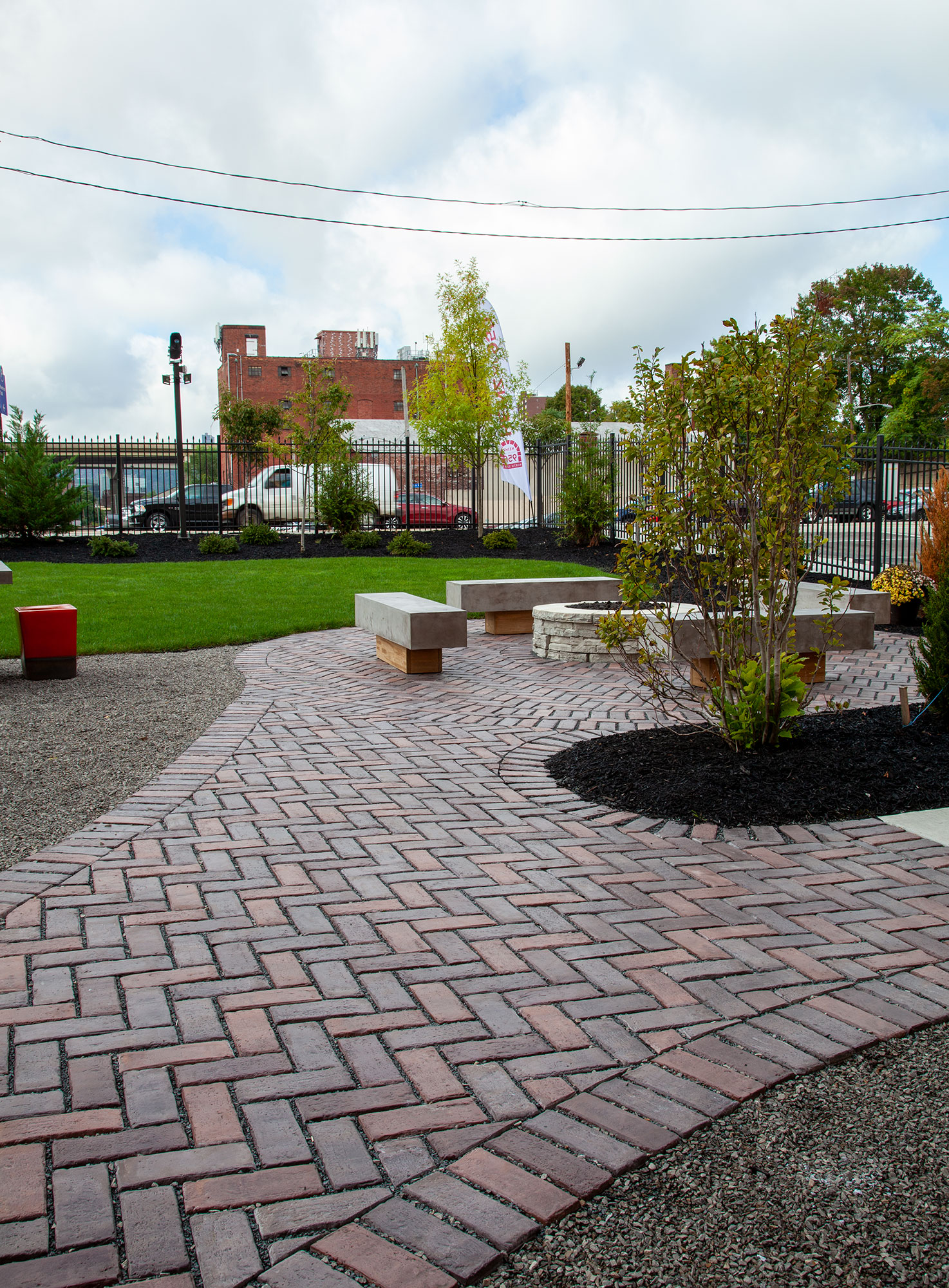 A circular fire pit and stone benches are surrounded by tri-colored Town Hall pavers, with nearby bushes adding subtle flecks of color.