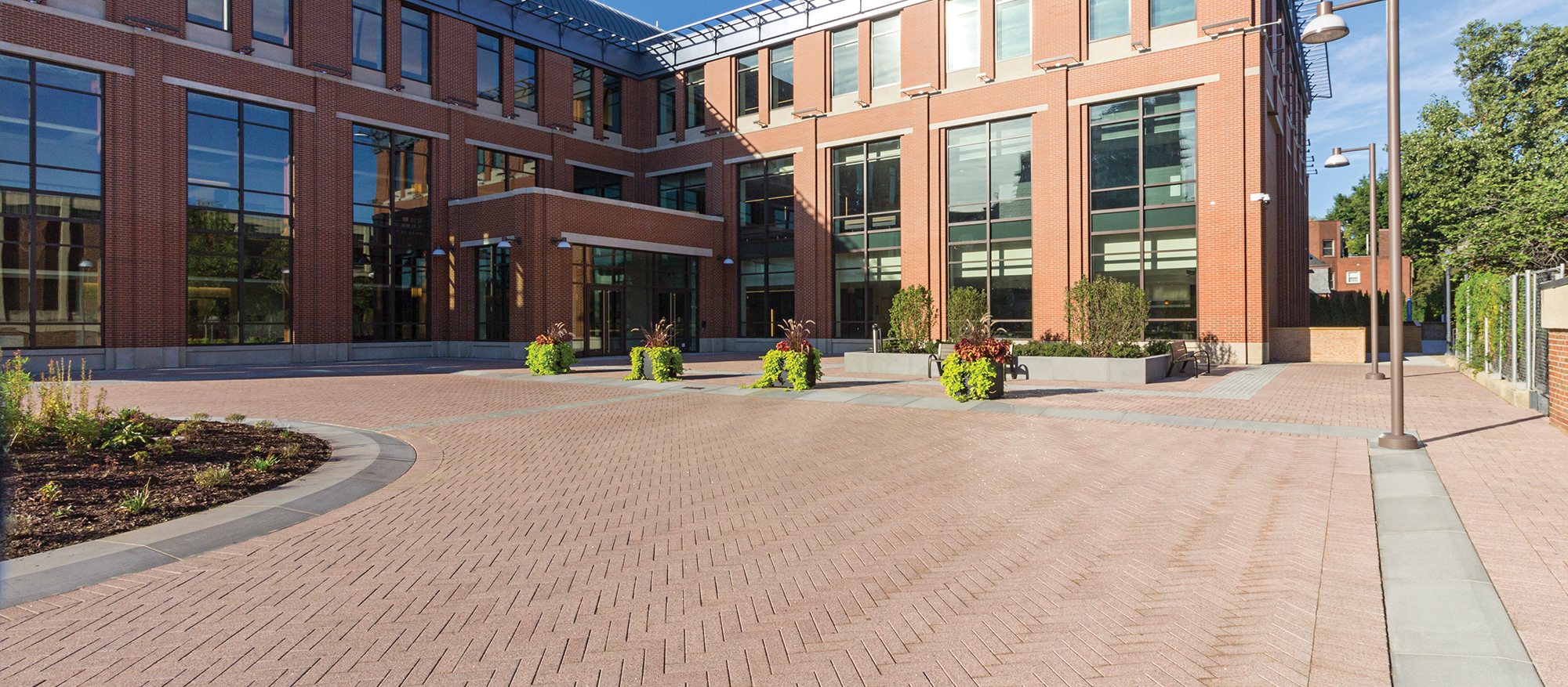 A courtyard paved with Promenade Plank in a herringbone pattern with grey accents, gardens and lighting leads to a red brick building.