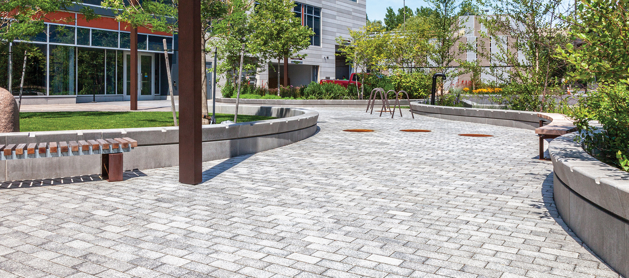 A rectangular building has a pedestrian area with Unilock Eco-Priora pavers in grey tones, with bike racks, benches, and landscaping.
