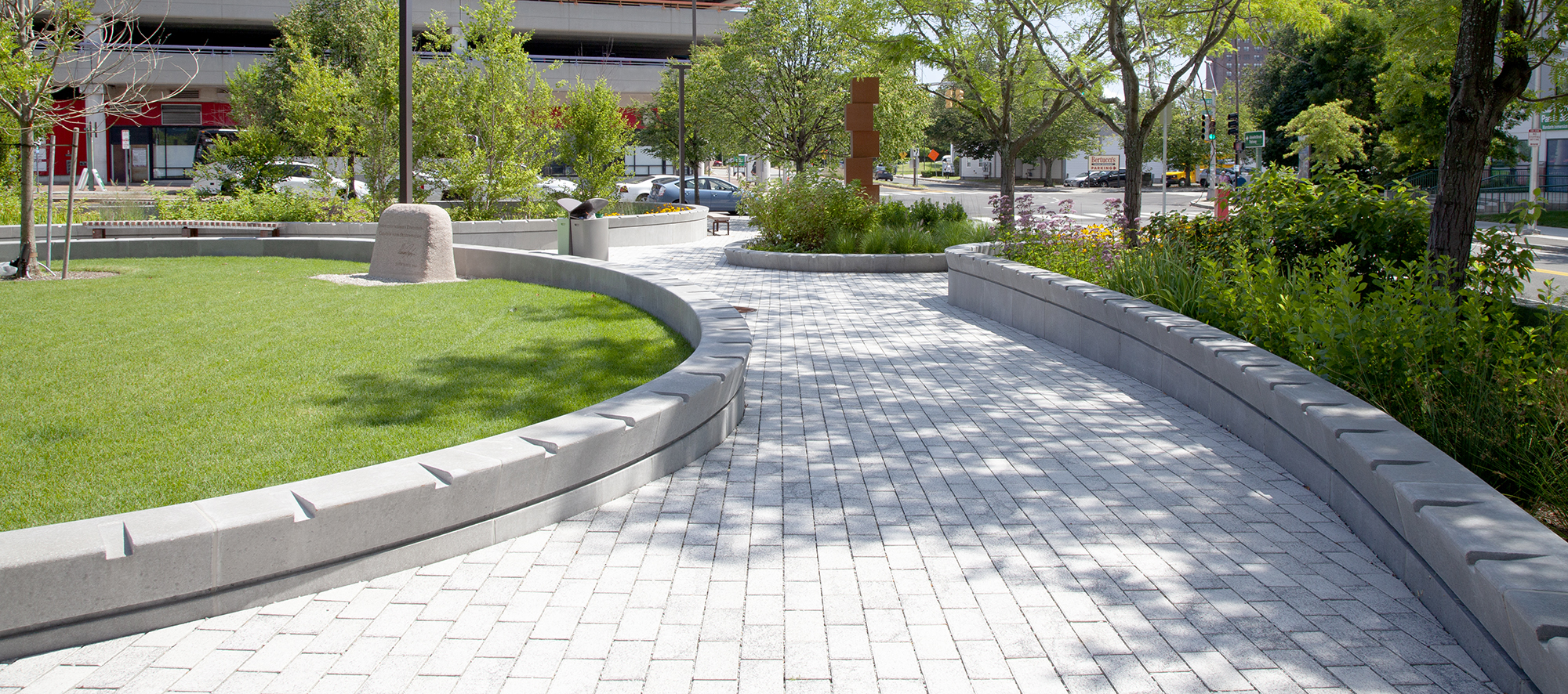 Unilock Eco-Priora pavers create a curved path bordered by raised gardens with a circular garden and block sculpture at the end.