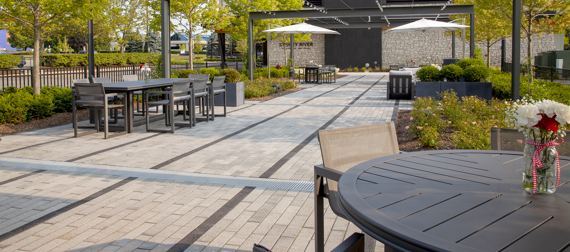 Two-toned Promenade Plank pavers delineate rows of walking spaces, with outdoor tables, chairs and umbrellas.