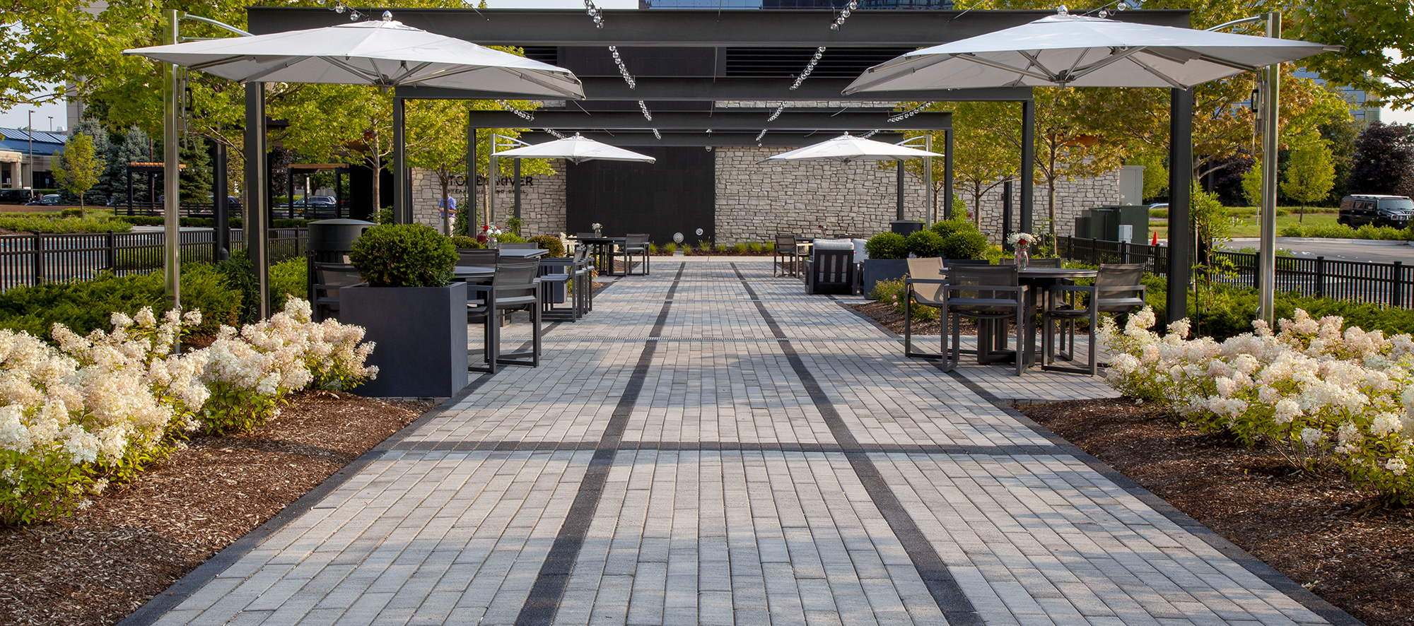 Promenade Plank pavers create a striped pattern in two tones under a pergola, with tables, chairs and landscaping.