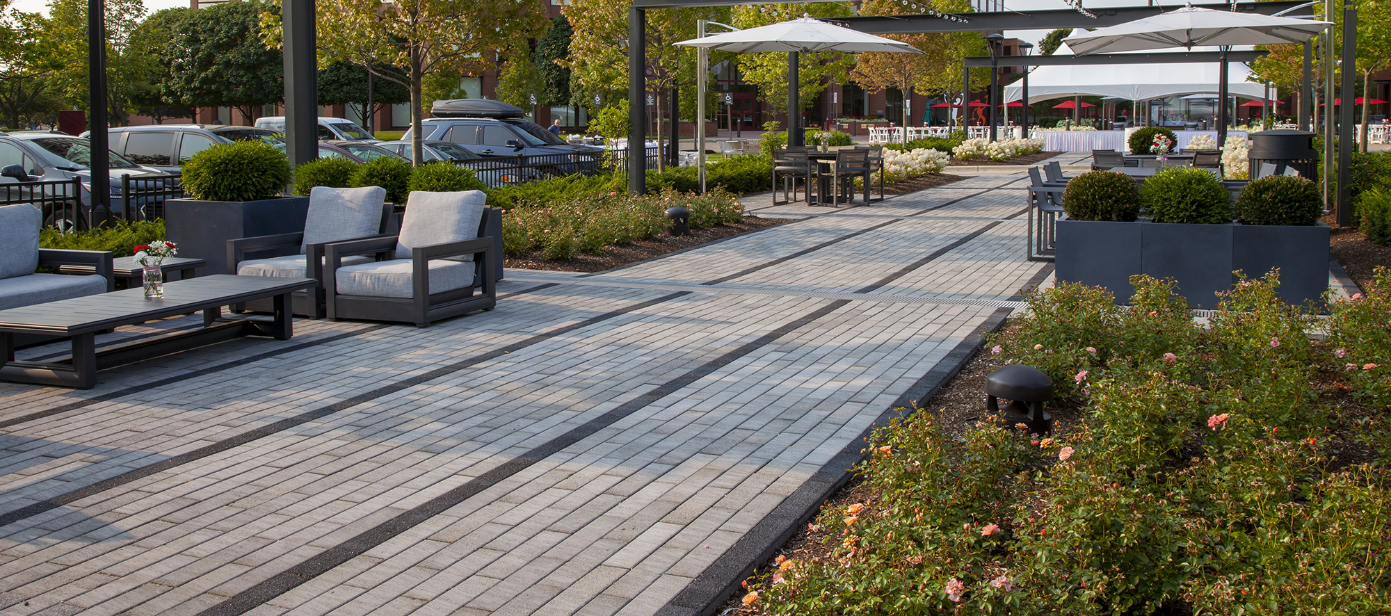 Two-toned Promenade Plank pavers delineate rows of walking spaces, with outdoor tables, chairs and umbrellas and landscaping on the sides.